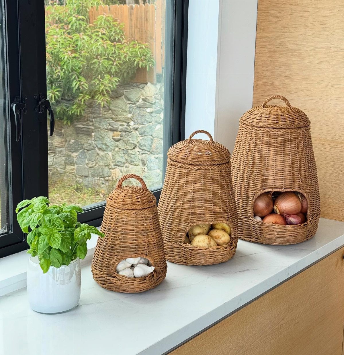 Three woven wicker storage baskets containing garlic, potatoes, and onions sit on a white kitchen countertop.