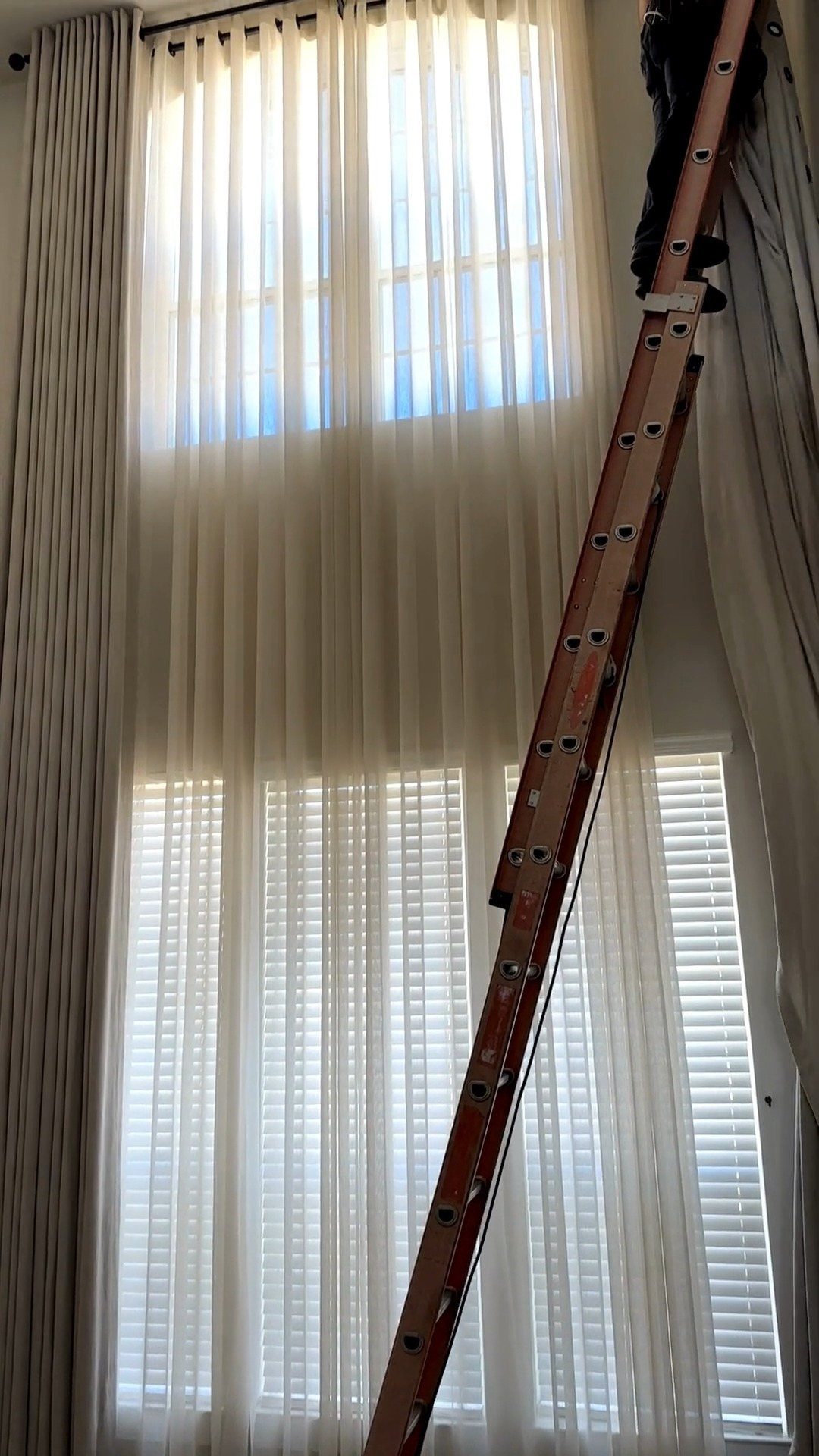 A wooden ladder leaning against tall, sheer white curtains covering a large window.