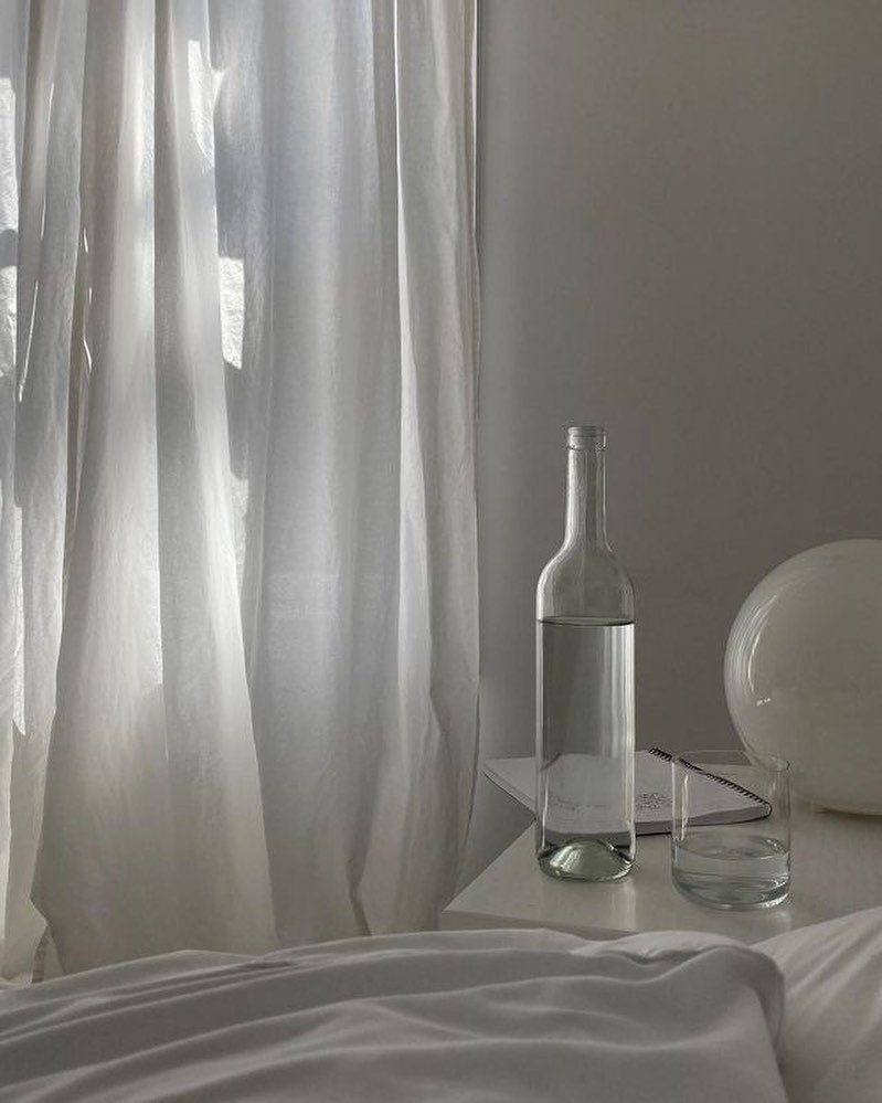 White curtains filter sunlight onto a minimalist bedside table with a glass bottle and water.