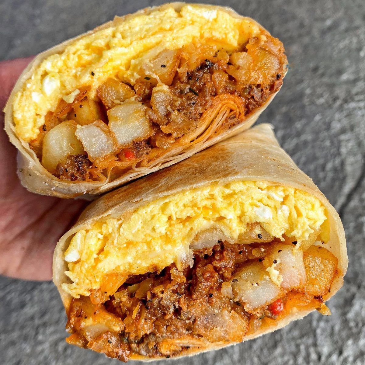 Two sliced breakfast burritos filled with eggs, meat, and potatoes held in a hand.