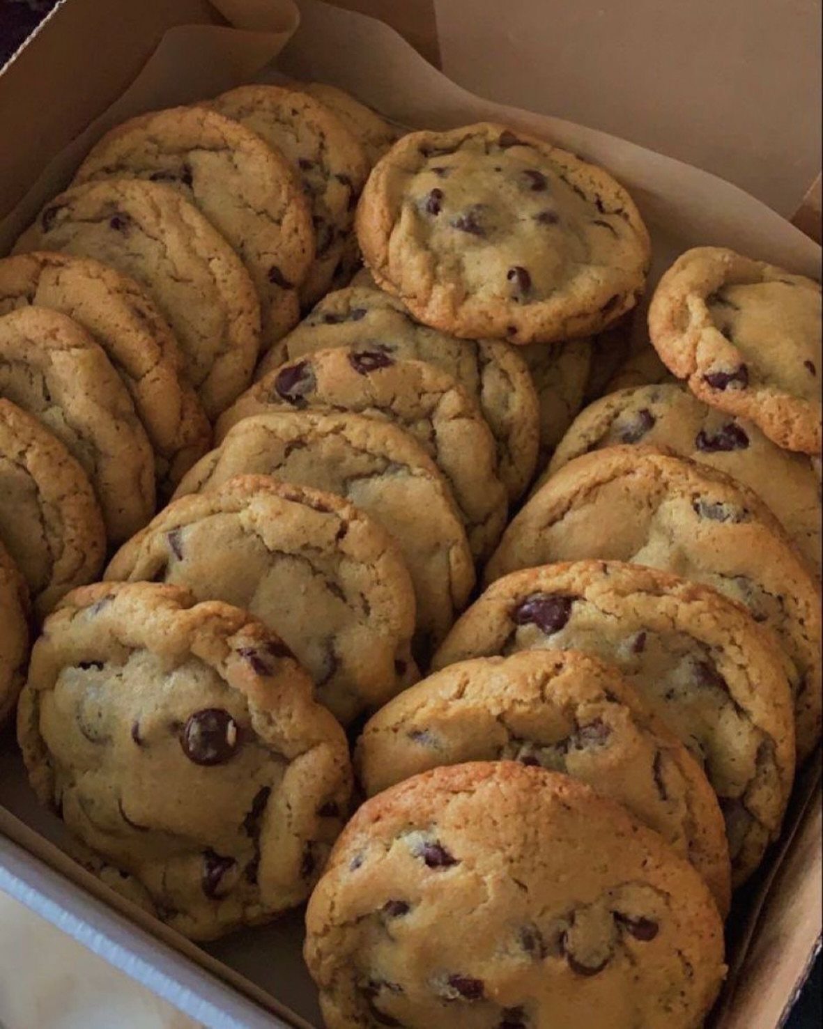 Many golden chocolate chip cookies piled inside a parchment-lined cardboard box.