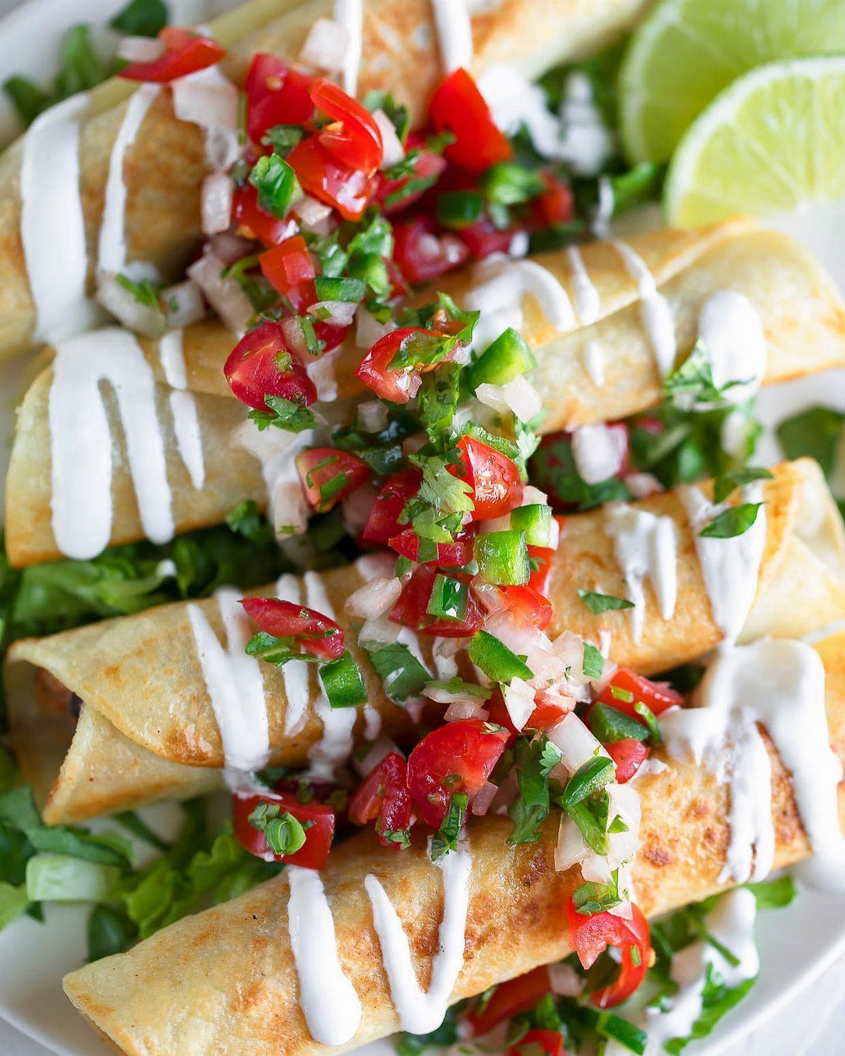 Golden taquitos topped with white crema, tomato salsa, and fresh cilantro served with lime wedges.
