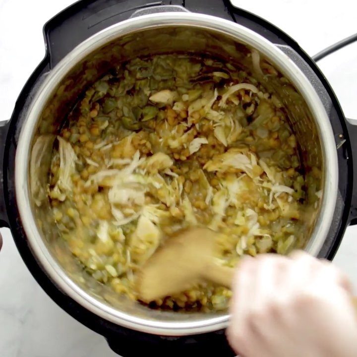 A person stirring a mixture of lentils and vegetables inside an electric pressure cooker with a wooden spoon.