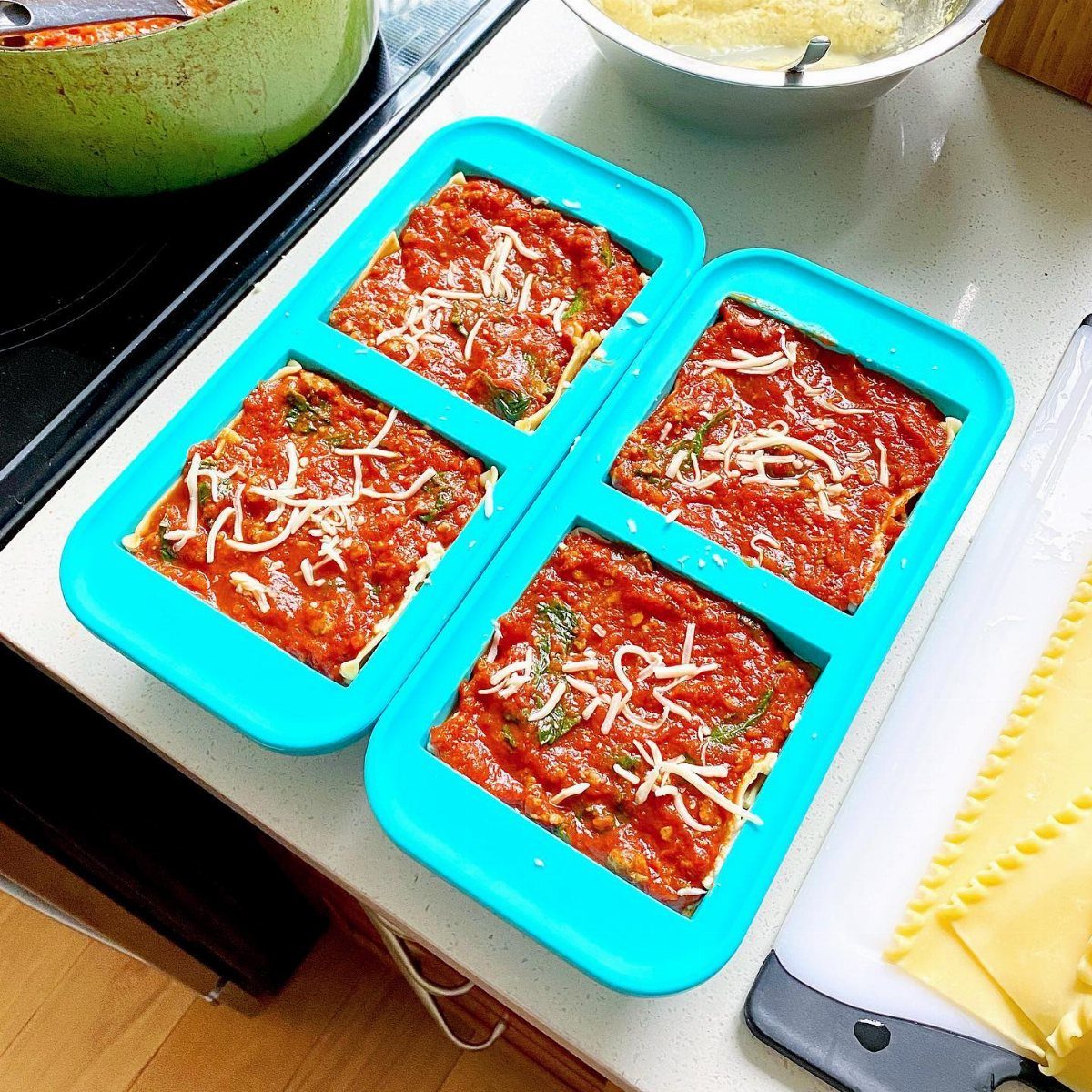 Two blue silicone molds filled with uncooked lasagna layers topped with red sauce and cheese.