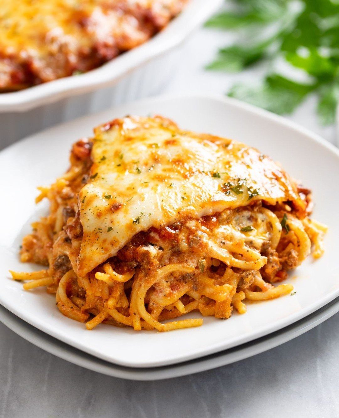 Baked spaghetti casserole with melted cheese served on a white plate with parsley in the background.