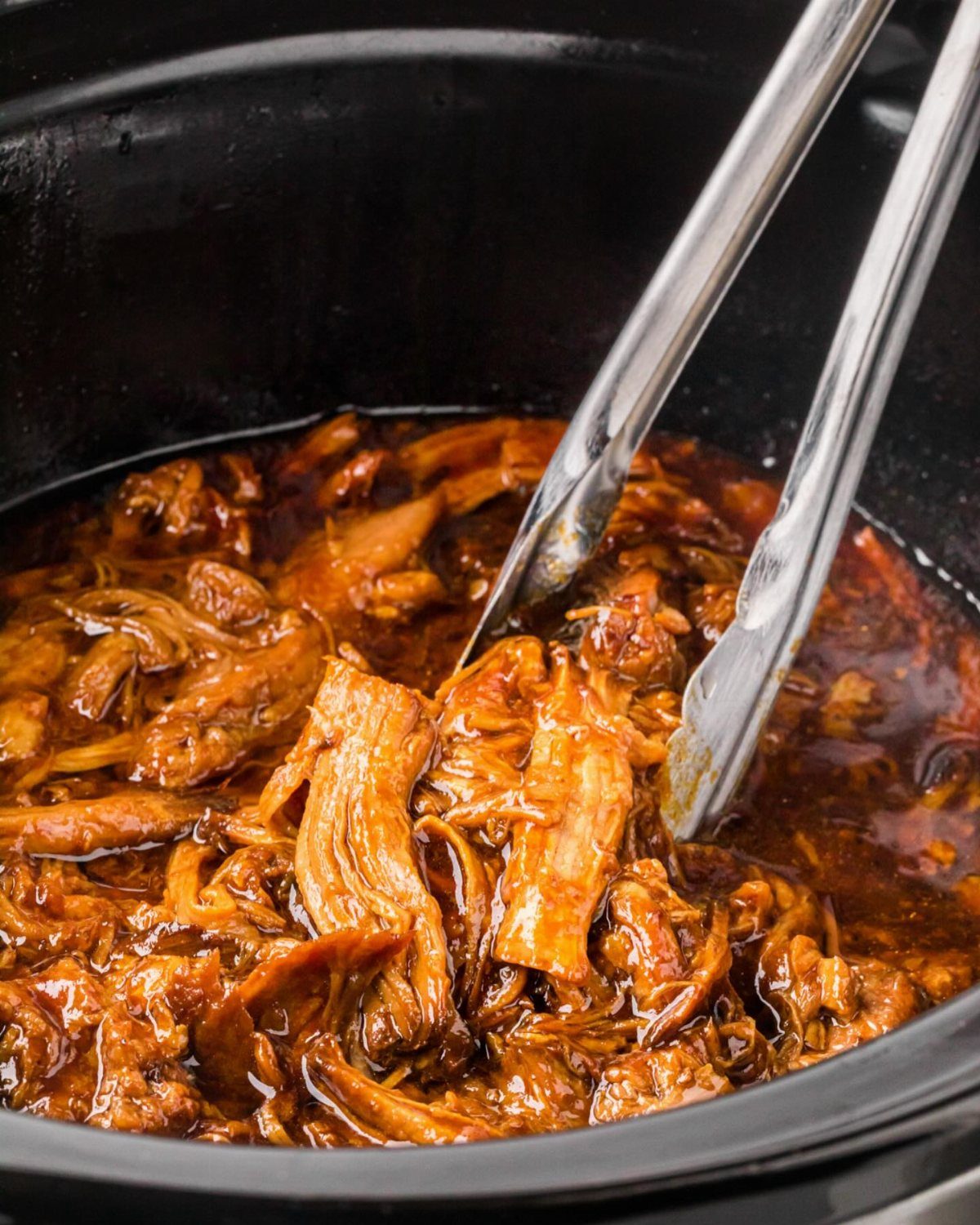 Metal tongs lifting shredded pulled pork from a dark sauce in a slow cooker.
