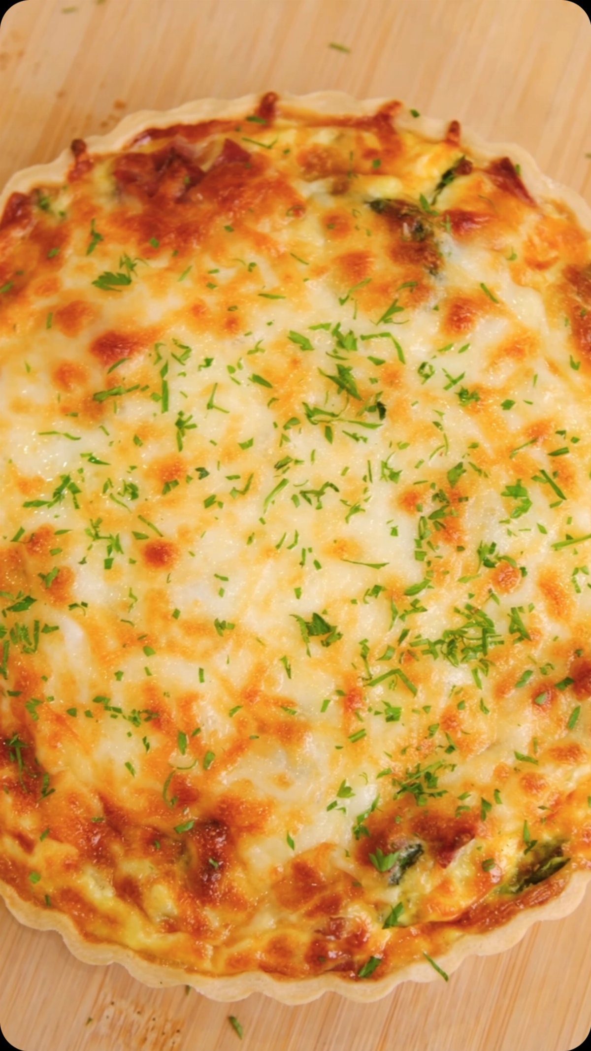 A golden baked quiche topped with melted cheese and fresh green herbs on a wooden board.