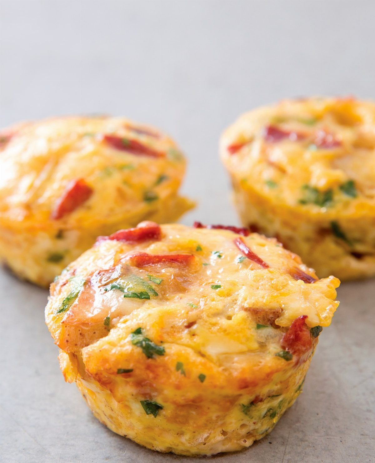Three savory egg muffins containing red peppers and green herbs on a grey background.