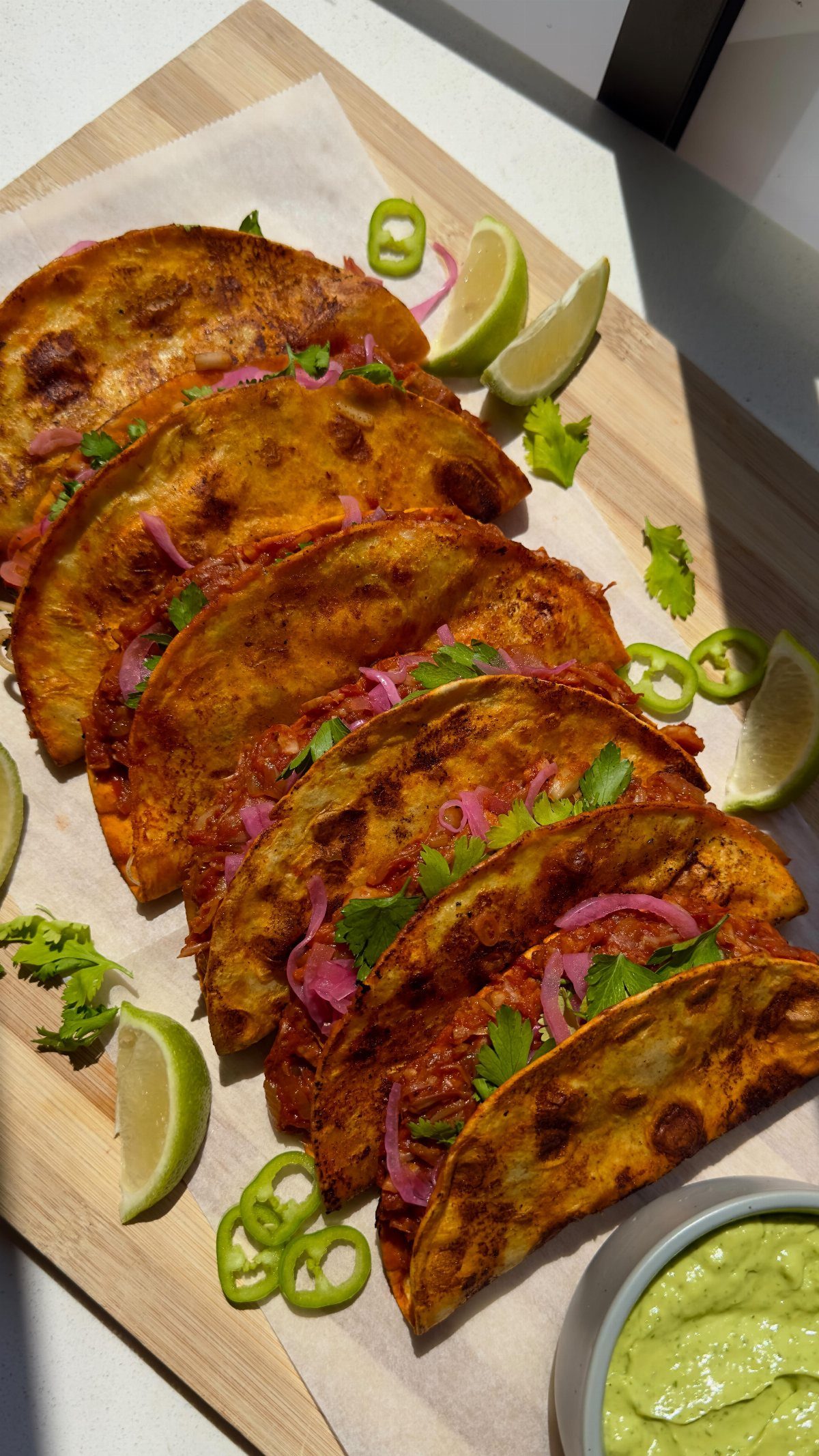 Crispy tacos topped with pickled onions and cilantro served on a wooden board with lime wedges.