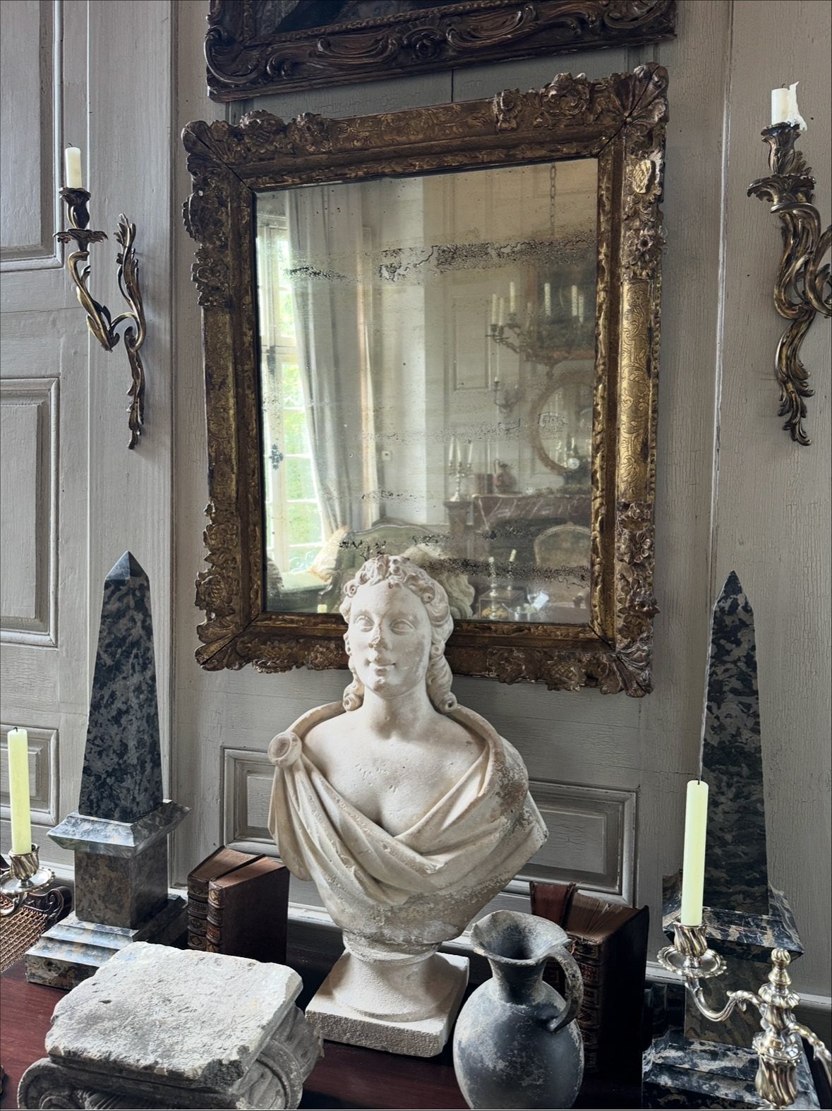 Ornate gold antique mirror hanging above a classical bust and stone obelisks on a mantel.
