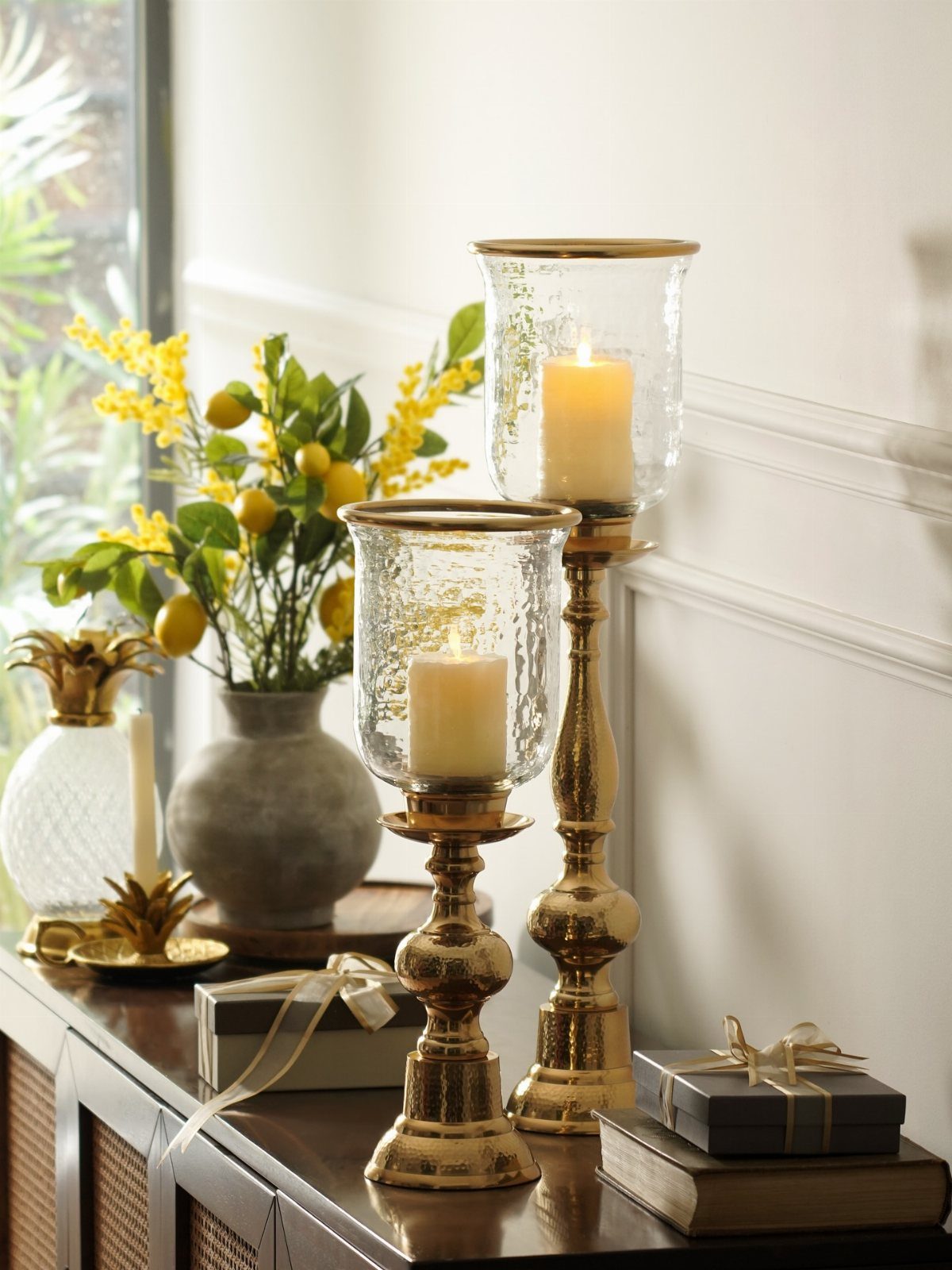 Gold glass candle holders with lit cream candles on a dark wooden sideboard.