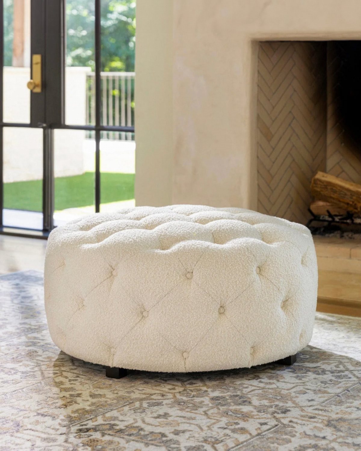 Cream tufted bouclé ottoman resting on a patterned rug in a bright living room.