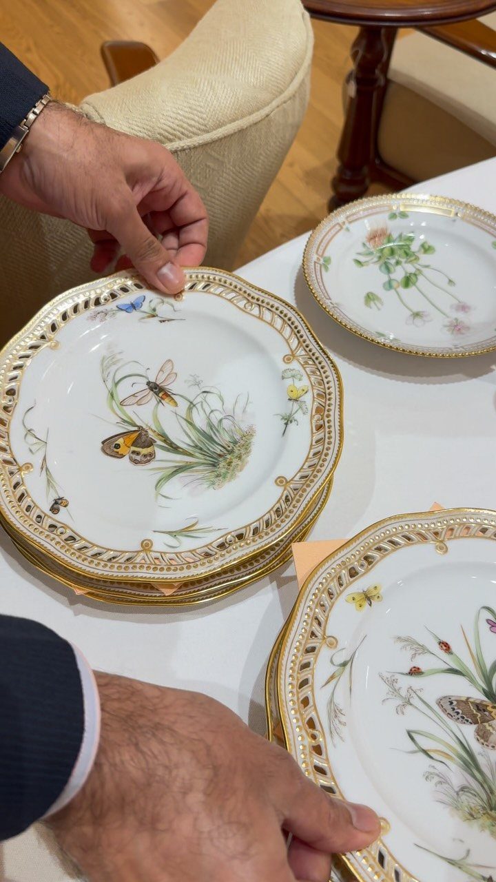 Hands arranging a stack of white and gold decorative plates with botanical insect illustrations.