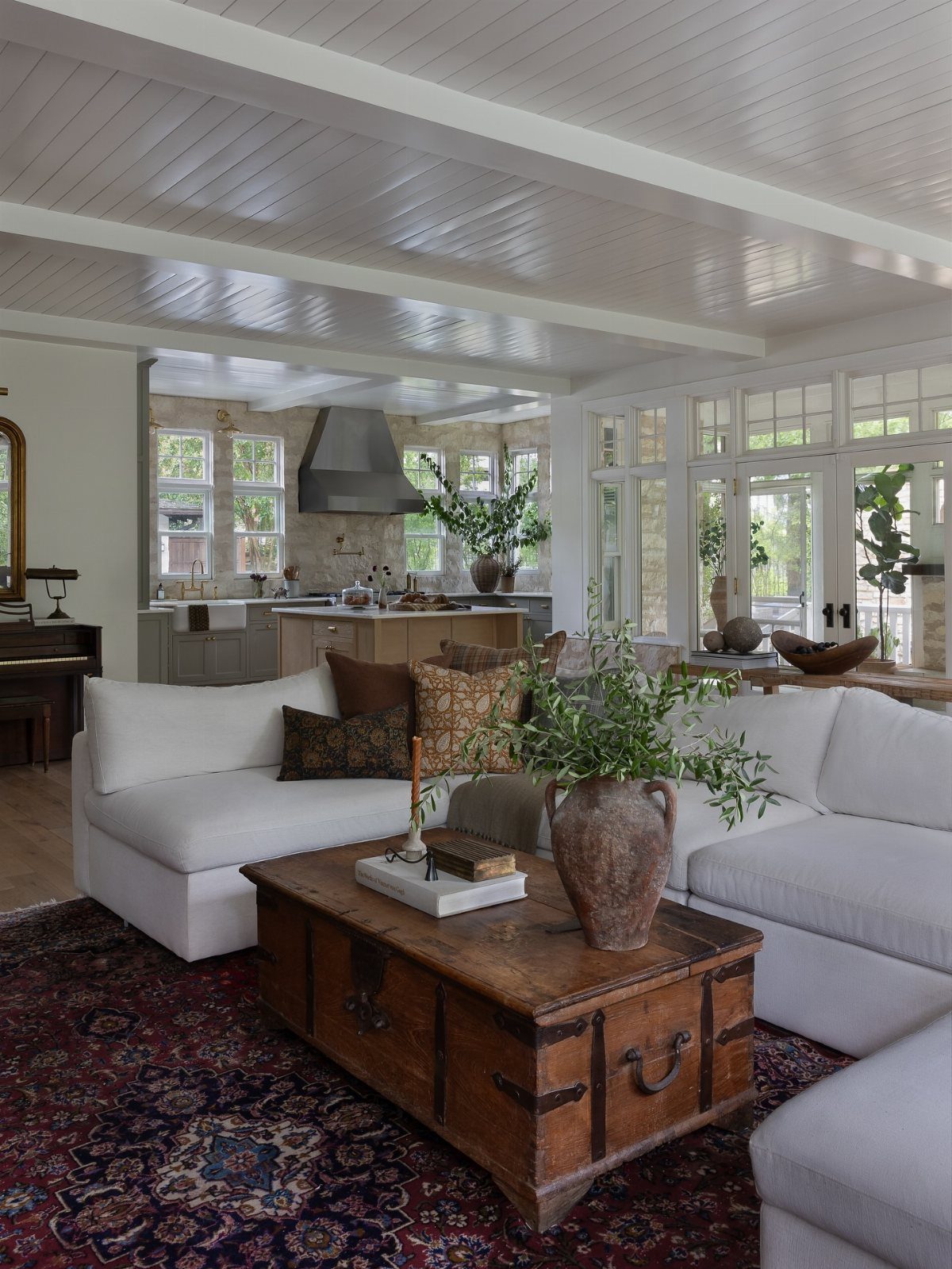 White sectional sofa with patterned pillows sits atop a dark patterned rug near a wooden trunk.