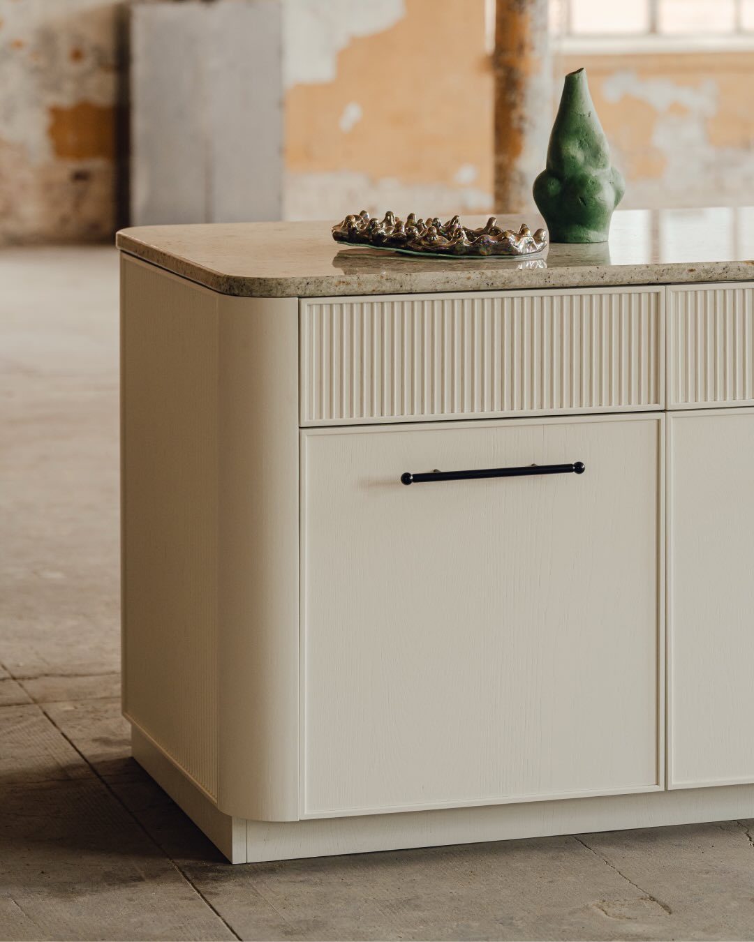 Cream fluted sideboard with a stone top, green vase, and decorative tray in a rustic room.