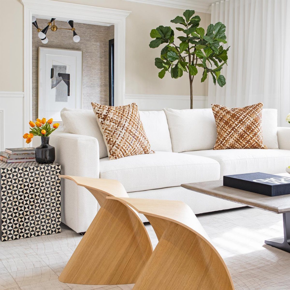 White sofa with patterned pillows, wooden sculptural tables, and a green plant in a bright room.