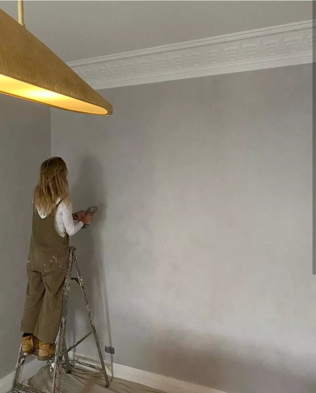 A person on a ladder painting a grey wall in a room with crown molding.