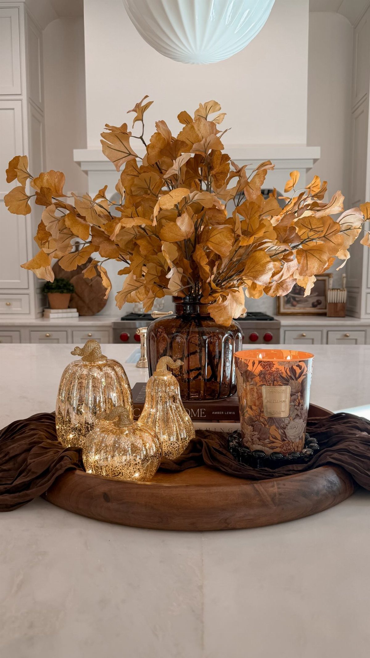 Dried golden leaves in a vase alongside gold pumpkins and a lit candle on a tray.