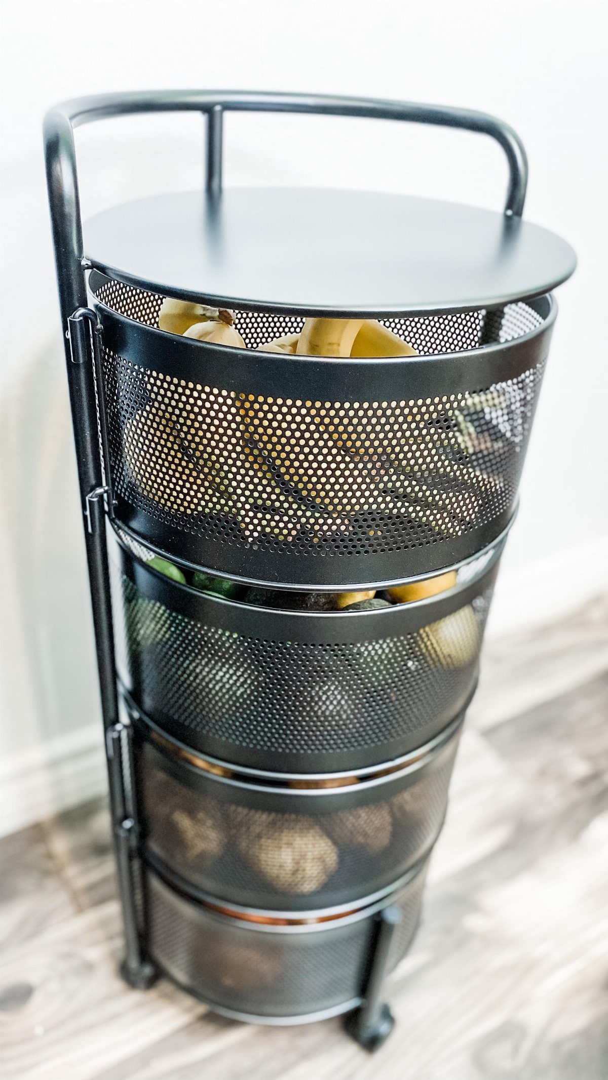 Black metal vertical tiered basket stand filled with various fruits and vegetables on a wooden floor.
