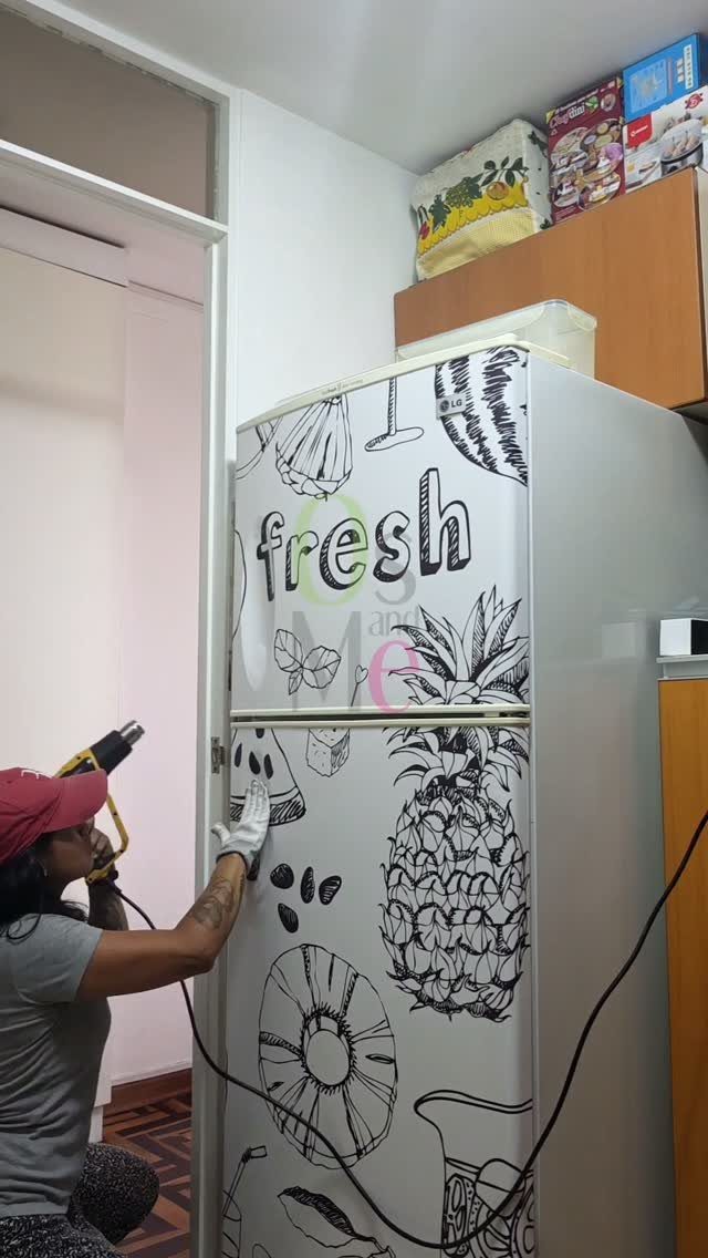 Person applying a fruit-patterned vinyl wrap to a white refrigerator door in a kitchen.