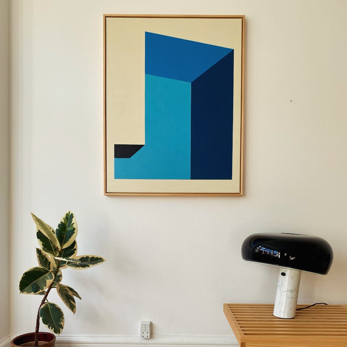 Abstract blue geometric art in a wooden frame hanging on a white wall above a modern lamp.