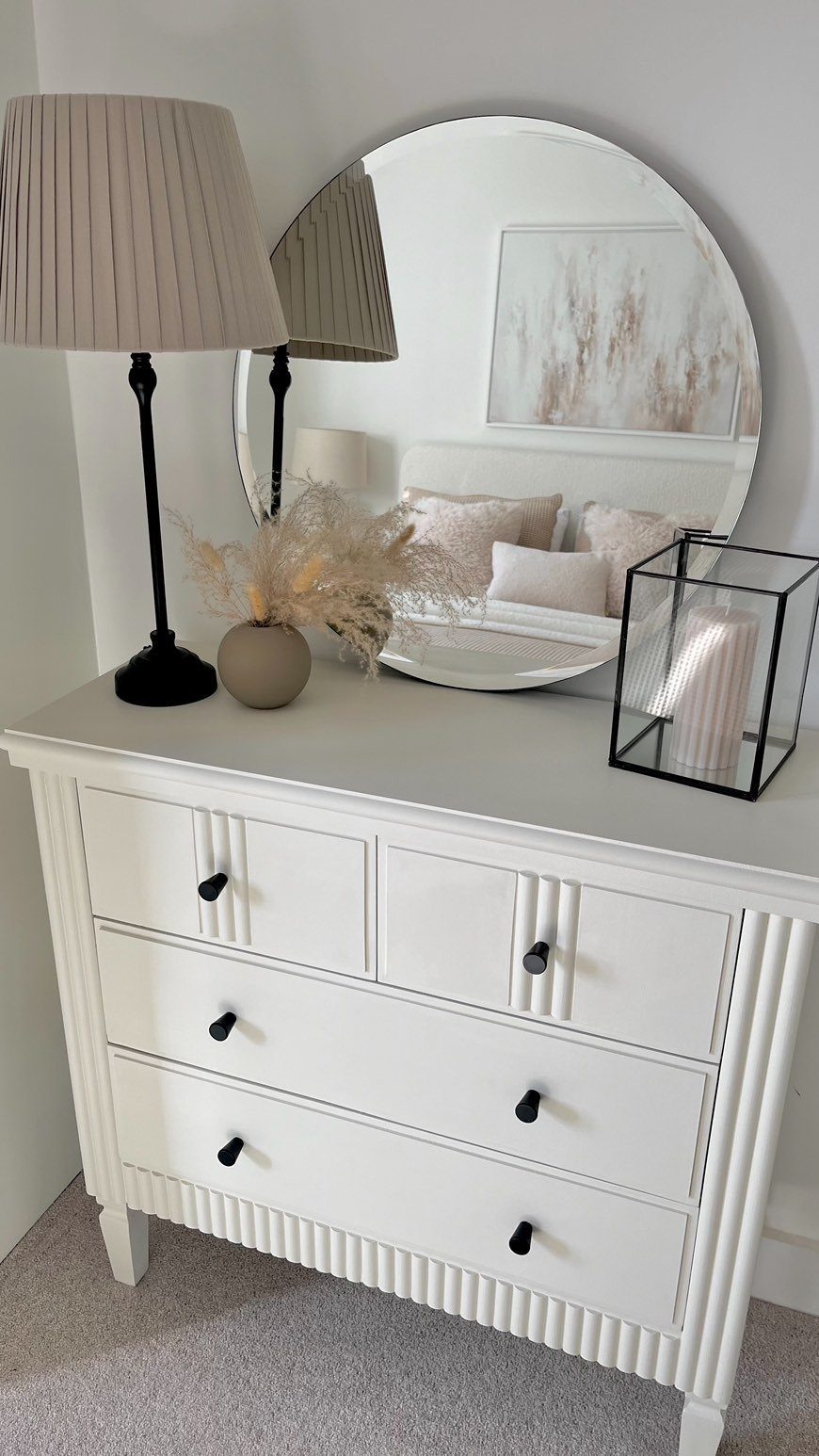White dresser decorated with a lamp, dried grass, and a large round mirror on the wall.