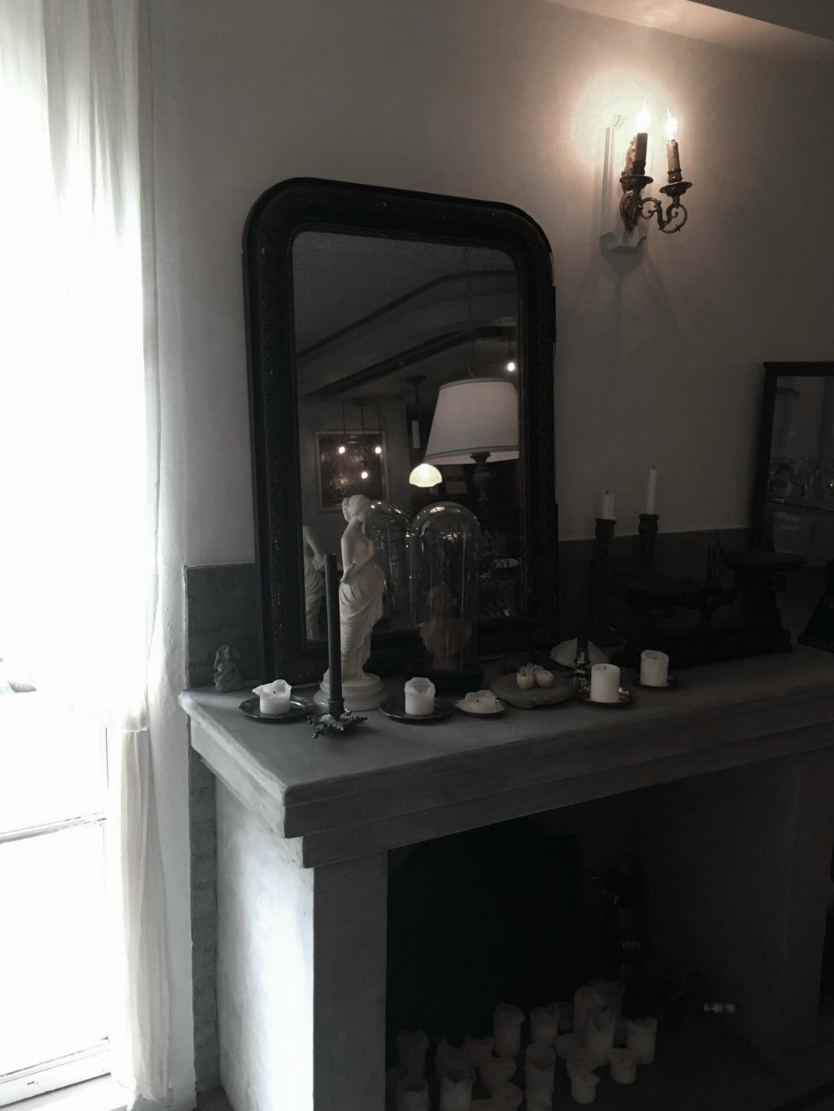 White candles and a glass cloche arranged on a stone mantelpiece near a dark mirror.