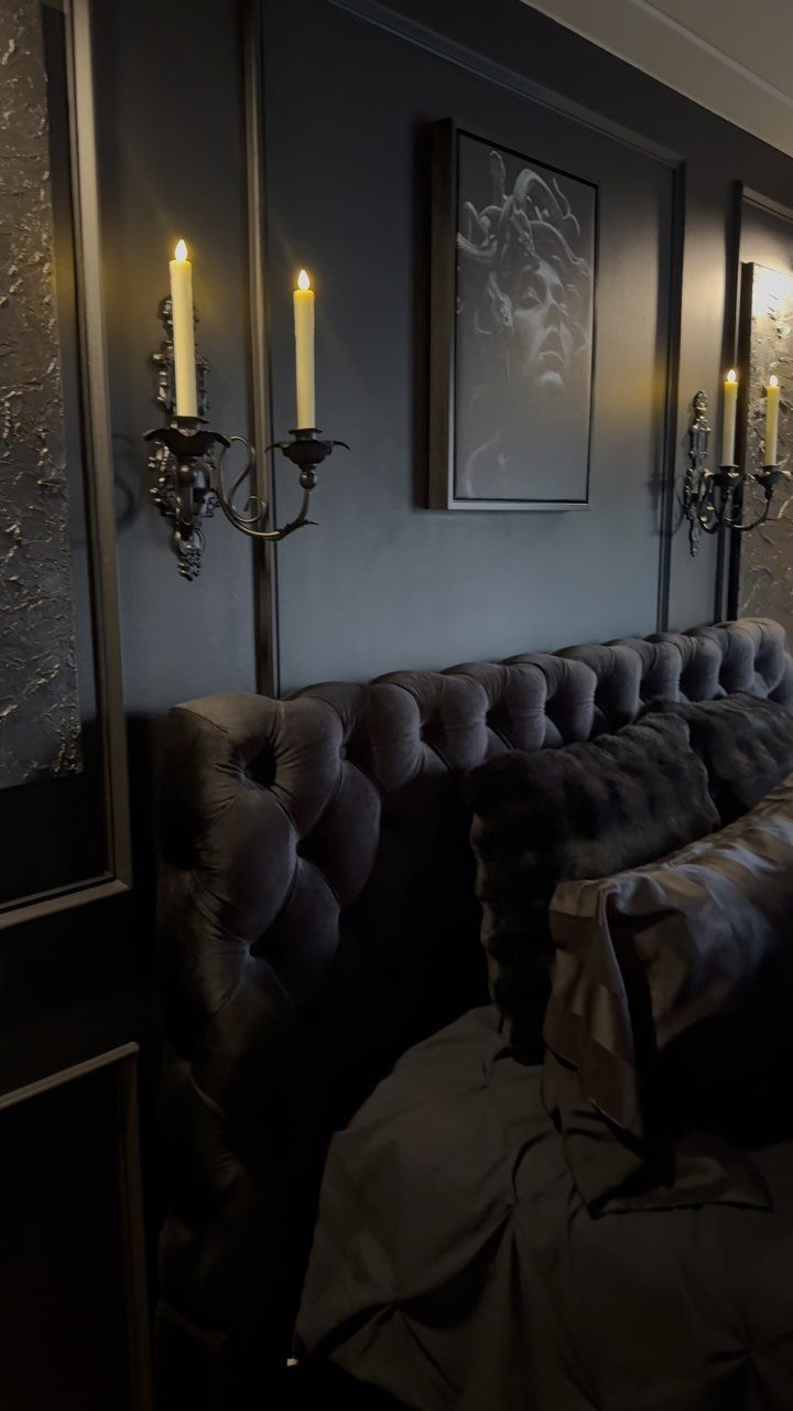 Tufted charcoal velvet headboard against black paneled walls with ornate candle sconces and framed art.