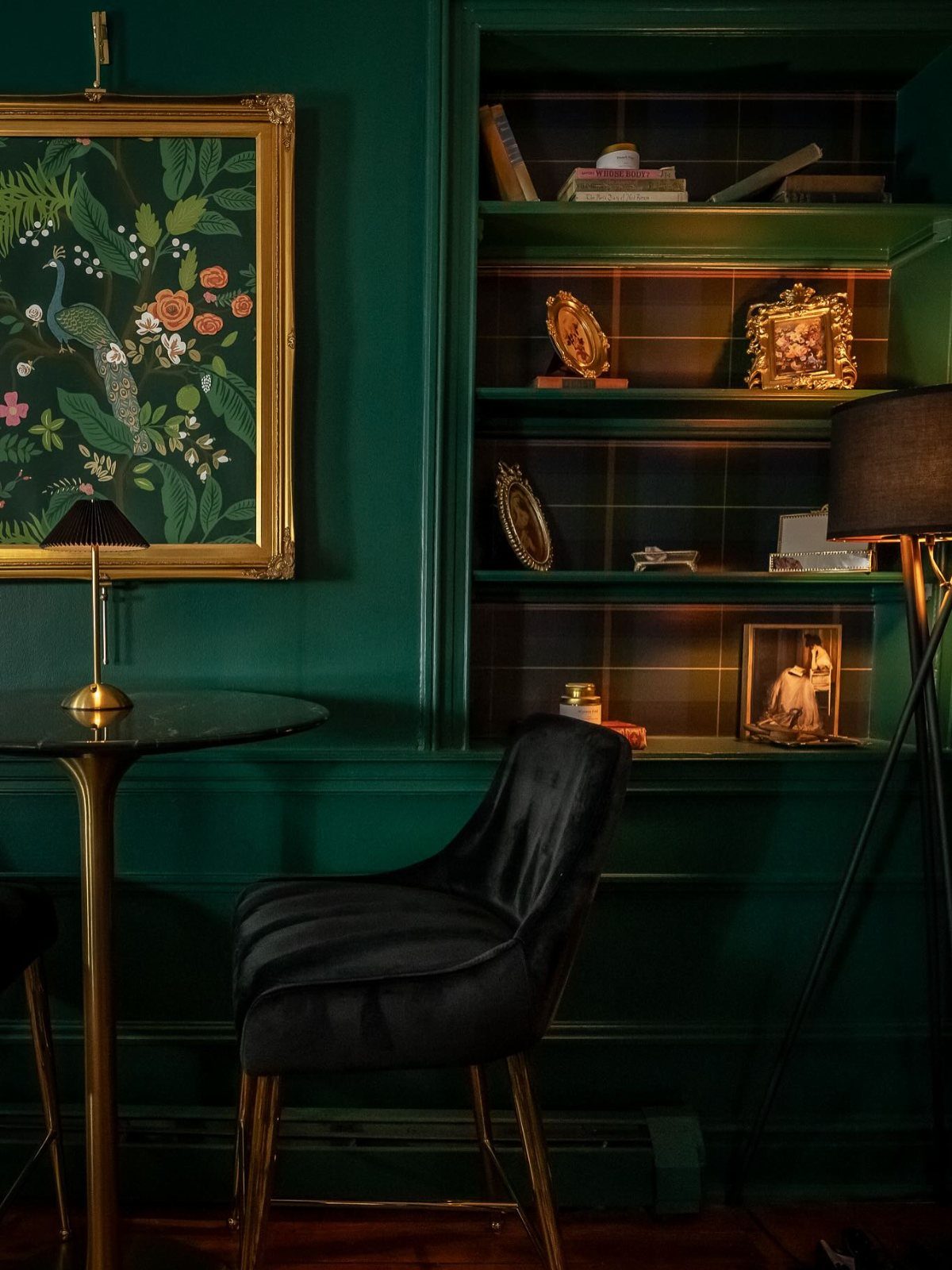 Dark emerald green walls in a room with a gold framed peacock painting and black velvet chair.