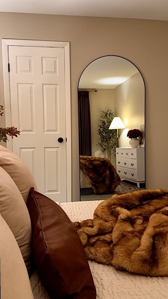 An arched full-length mirror reflecting a bedroom setup with a dresser and lamp.