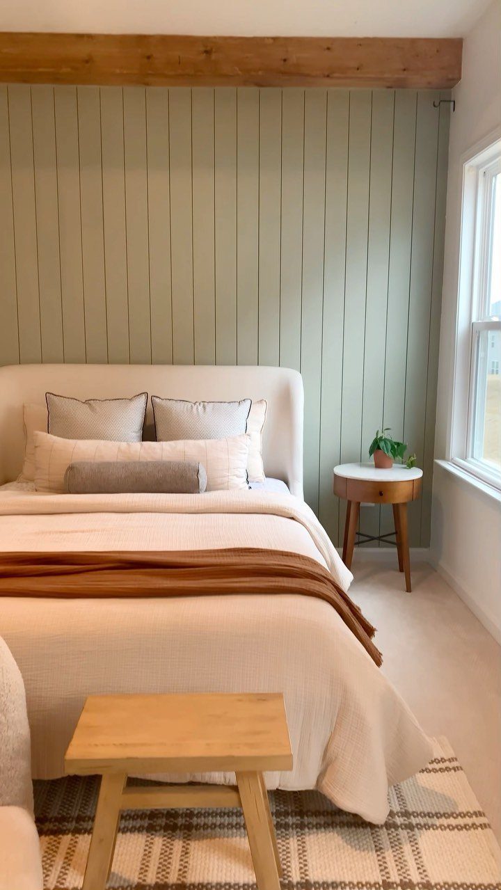 A neatly made bed with various neutral toned decorative pillows and linens in a sage green room.