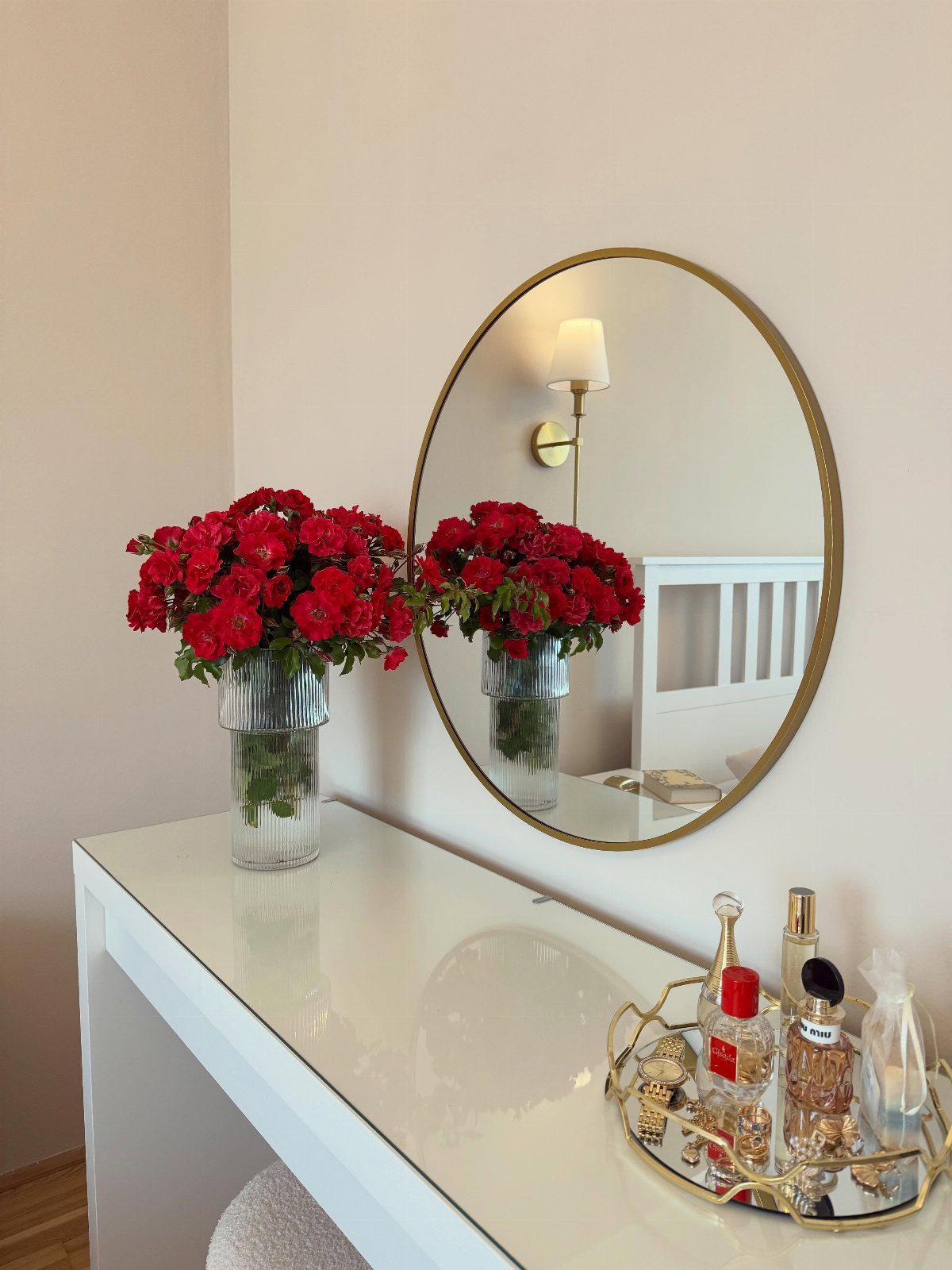 Red flowers in a glass vase on a white vanity next to a round mirror.