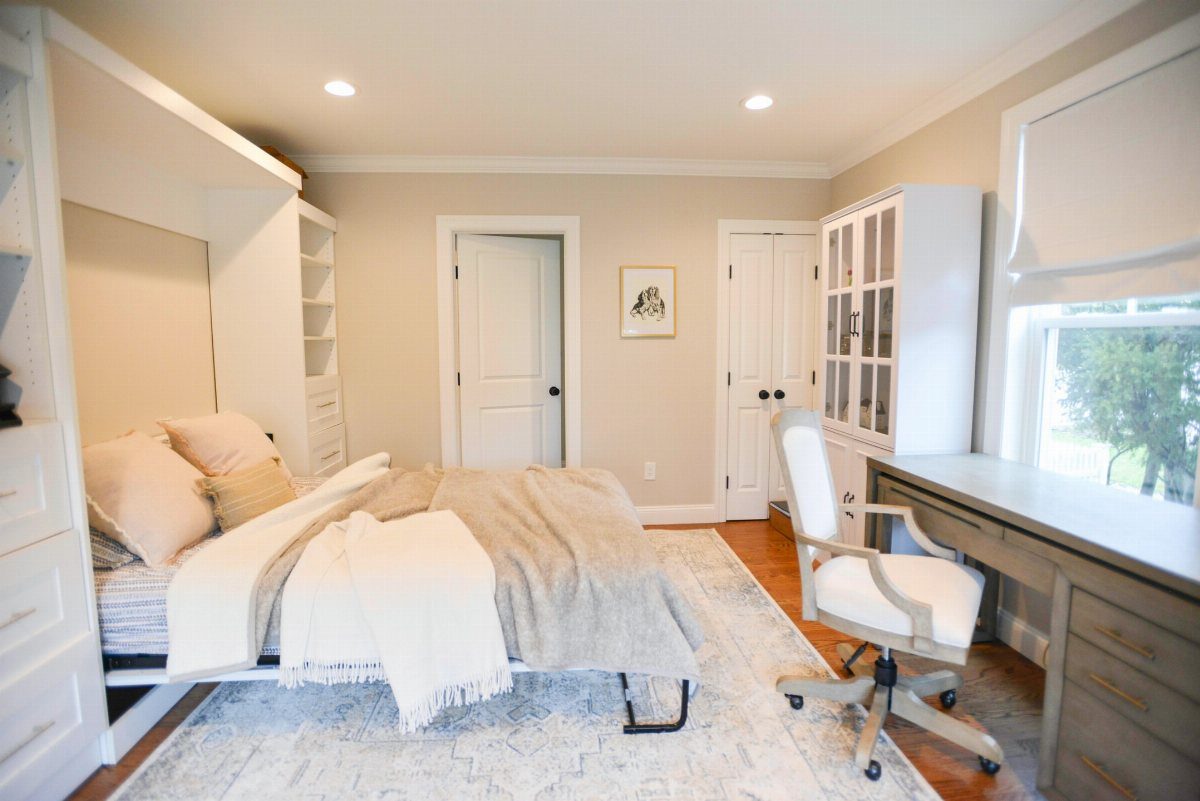 A wooden desk with a white office chair sits next to a bed in a bright room.