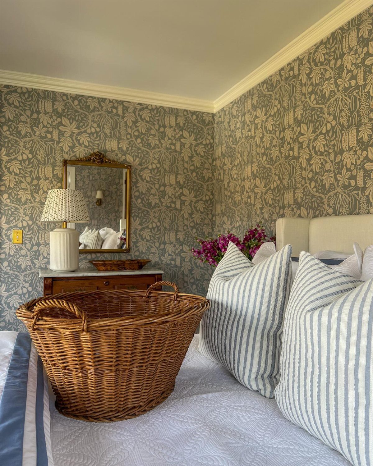 Floral patterned wallpaper covers the walls of a bedroom with a bed and wicker basket.