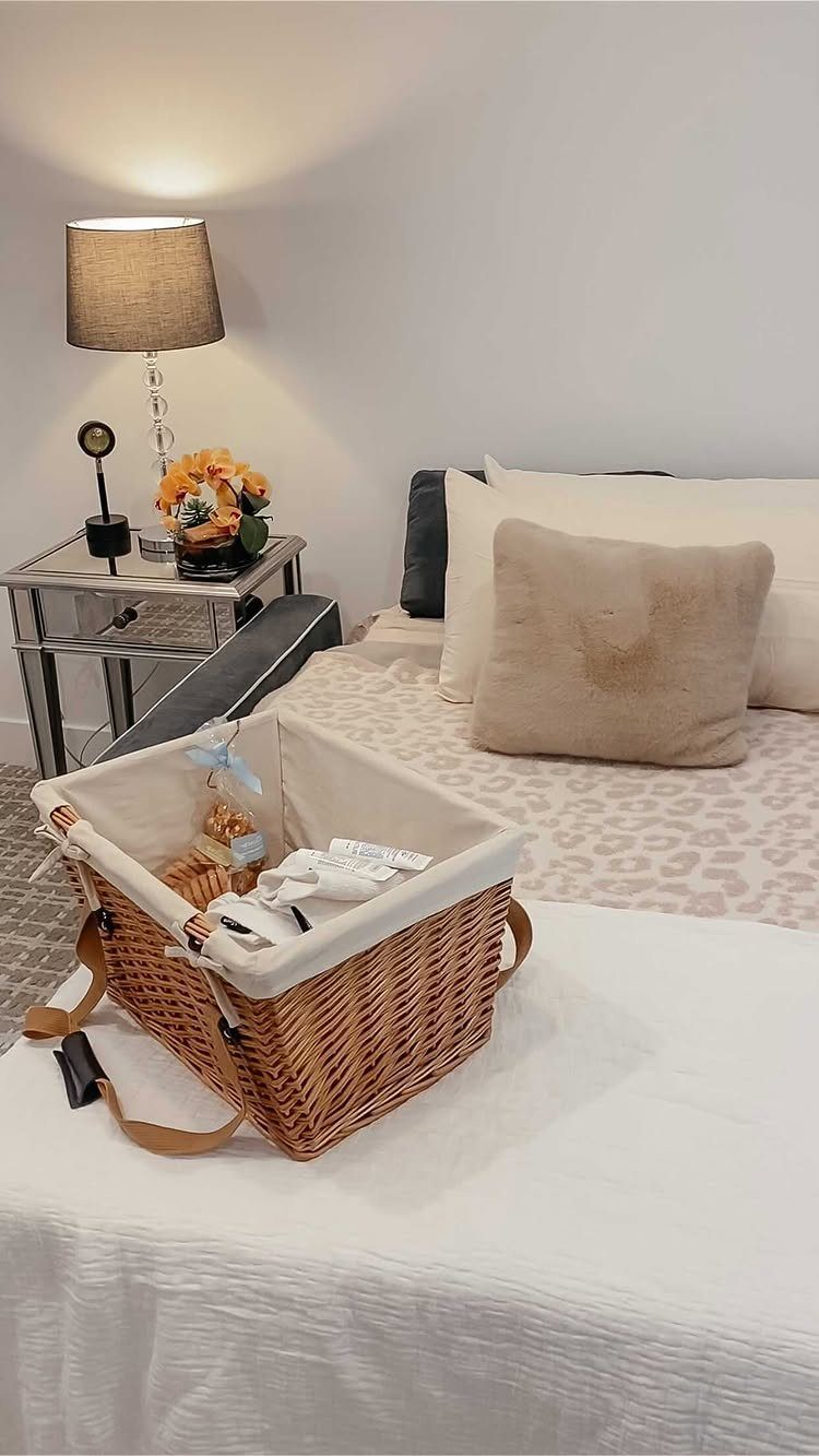 A wicker welcome basket with white fabric lining sits on a white table in a bedroom.
