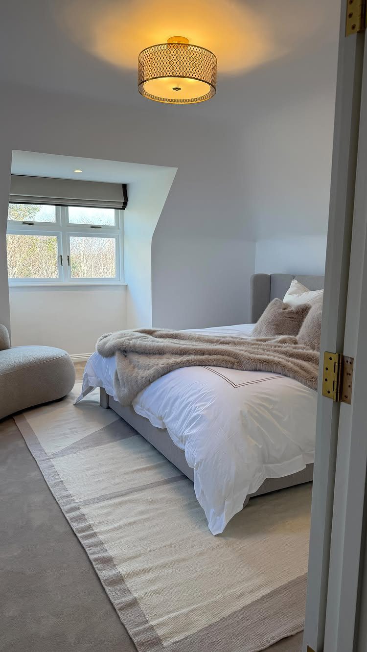 A bright guest bedroom with white bedding, a grey armchair, and soft neutral tones.