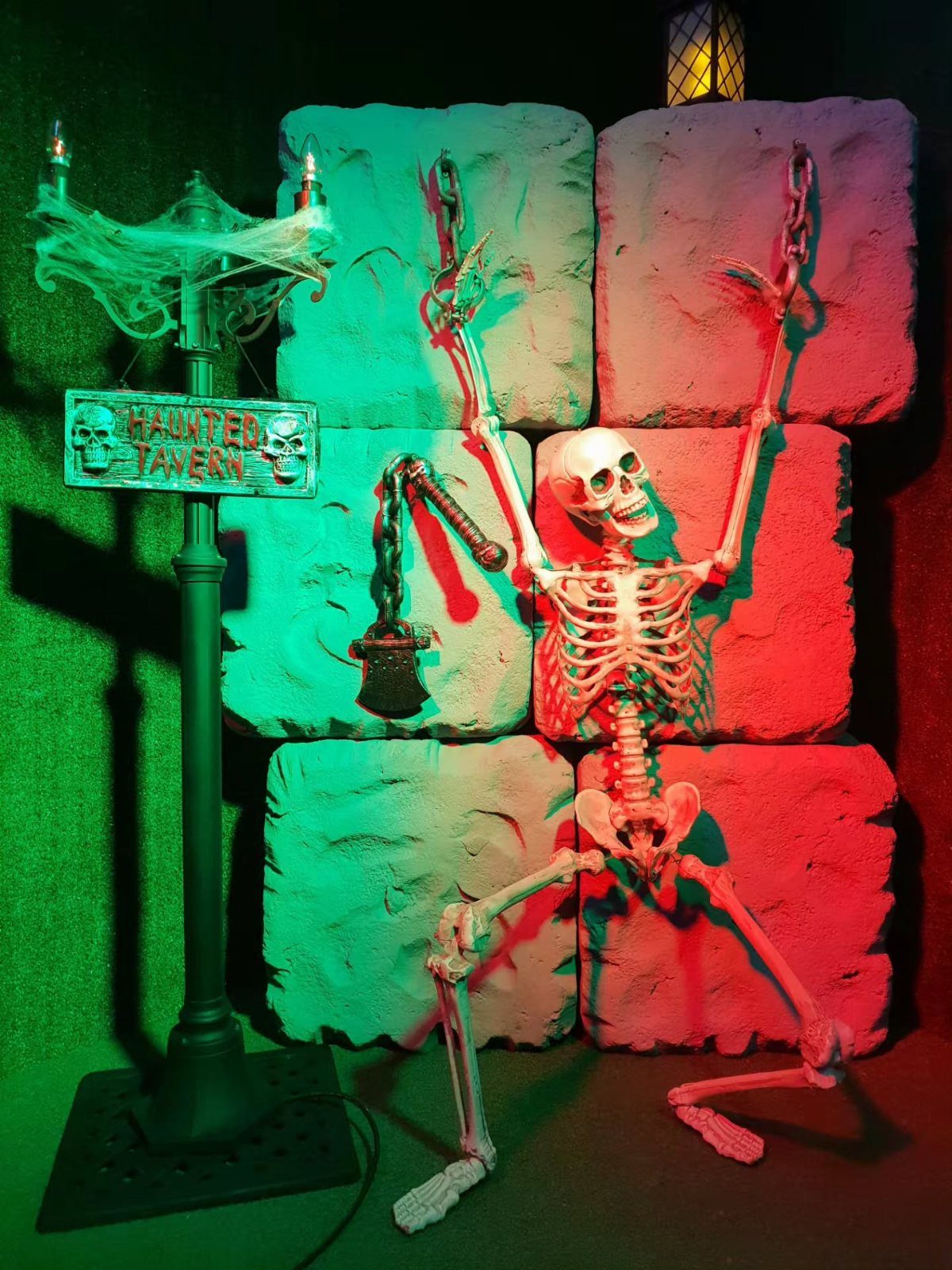 A skeleton chained to a stone wall next to a haunted tavern sign under red and green lights.