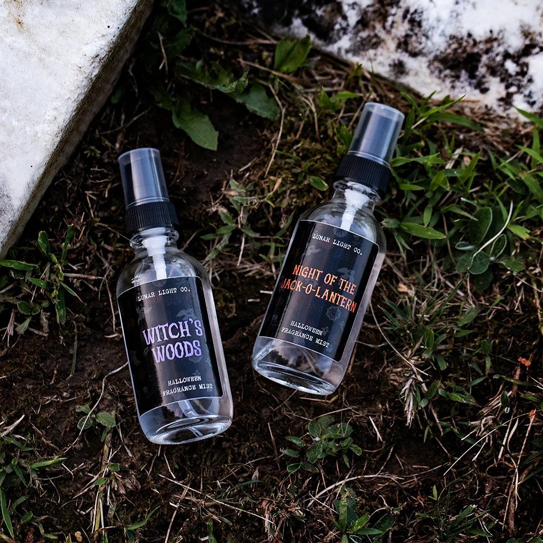 Two Halloween fragrance mist spray bottles resting on dark soil and green grass.