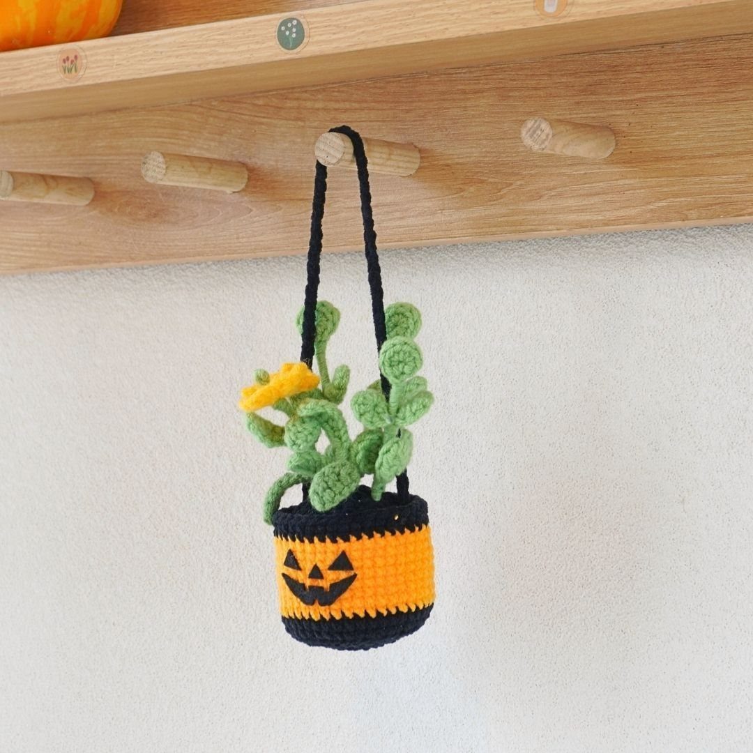 Crochet jack-o'-lantern hanging planter with a green plant against a white wall.