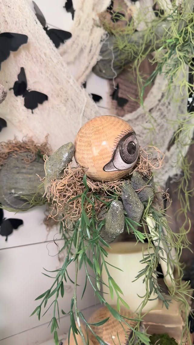 A large eyeball ornament resting in green moss and vines with white gauzy fabric.