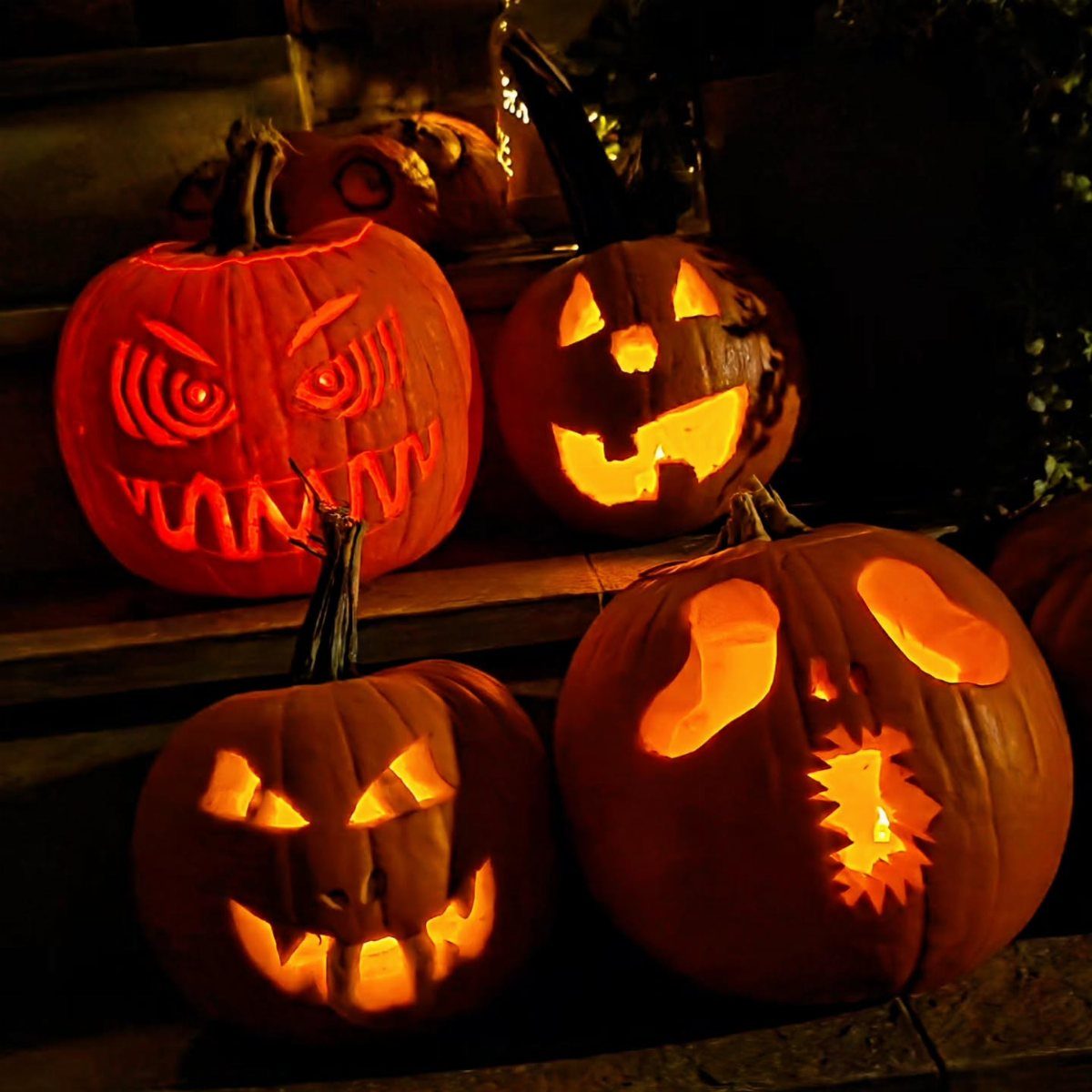 Four glowing carved jack-o'-lanterns arranged on dark wooden steps at night.