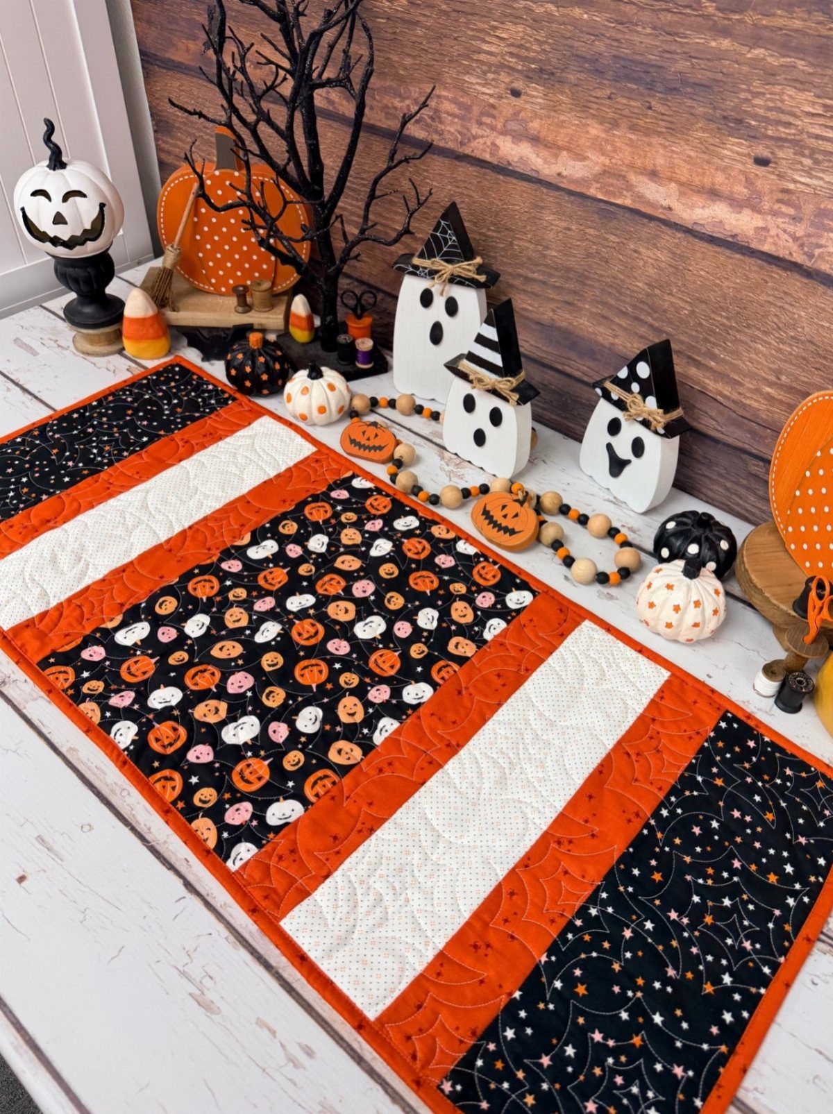 A quilted Halloween table runner with orange, black, and white patterns surrounded by spooky decor.