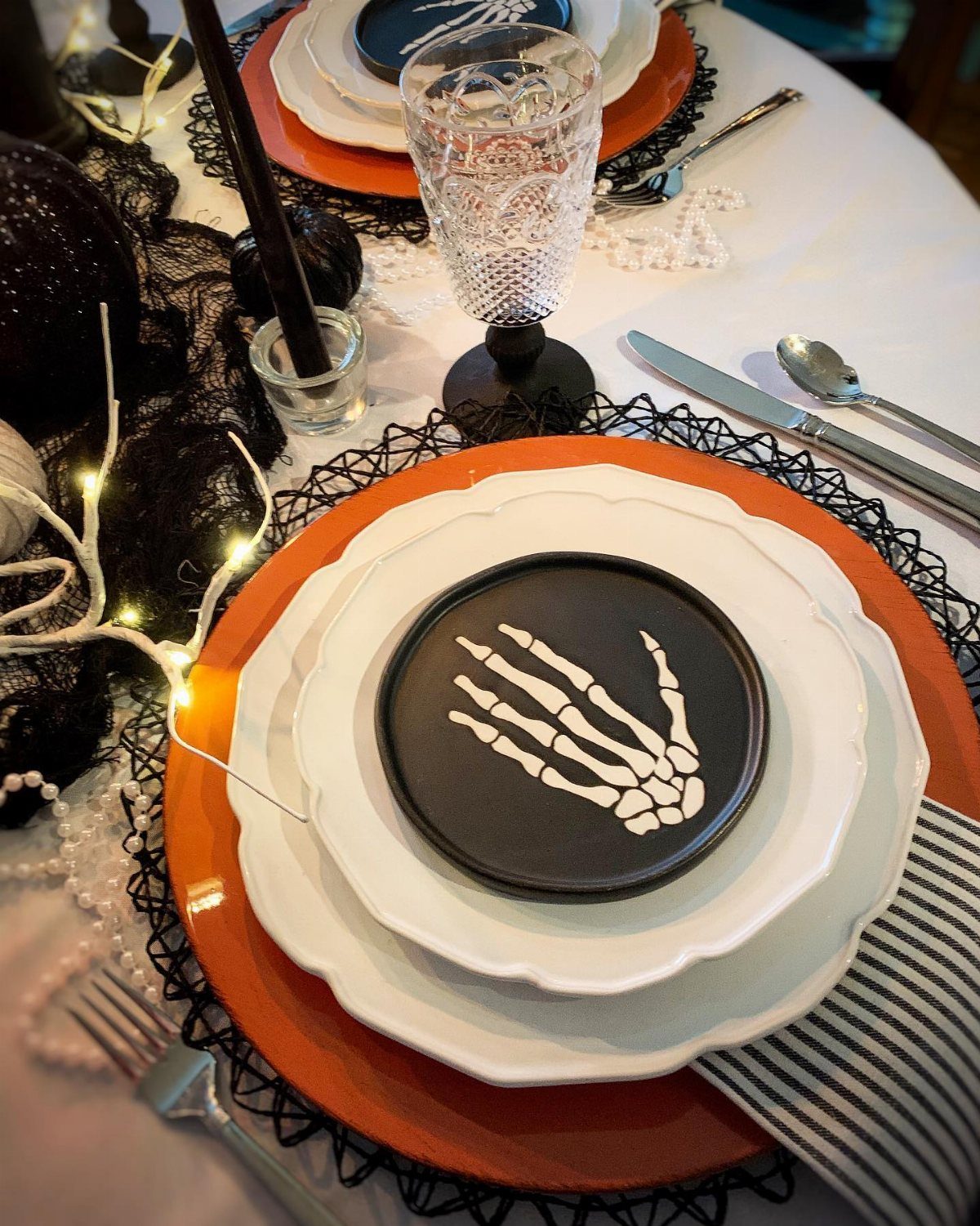 Halloween table setting with orange plates, skeleton hand dishware, and decorative string lights on a white cloth.