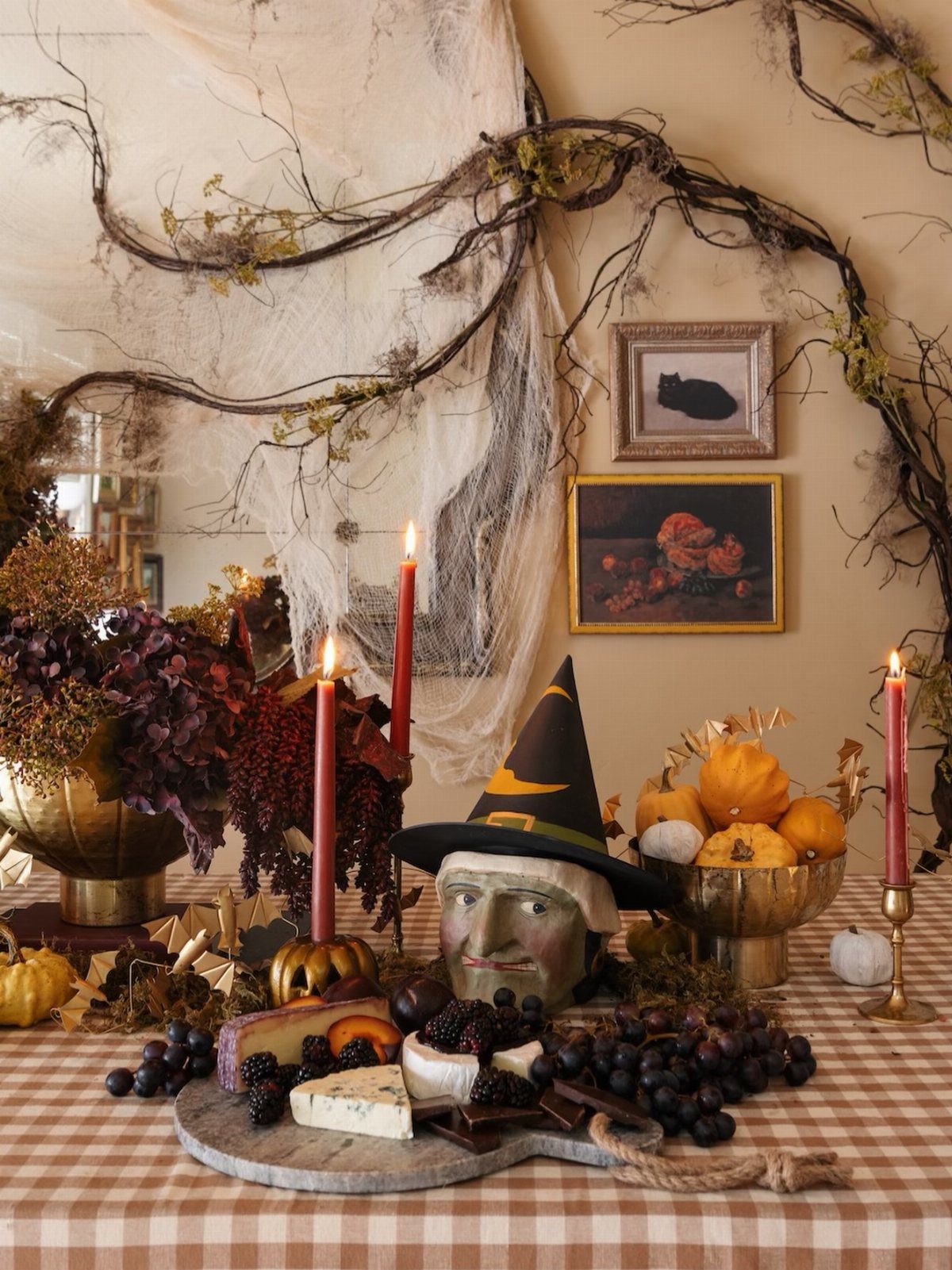 Tall red taper candles flickering amidst a Halloween centerpiece with a witch hat, pumpkins, and dark fruit.