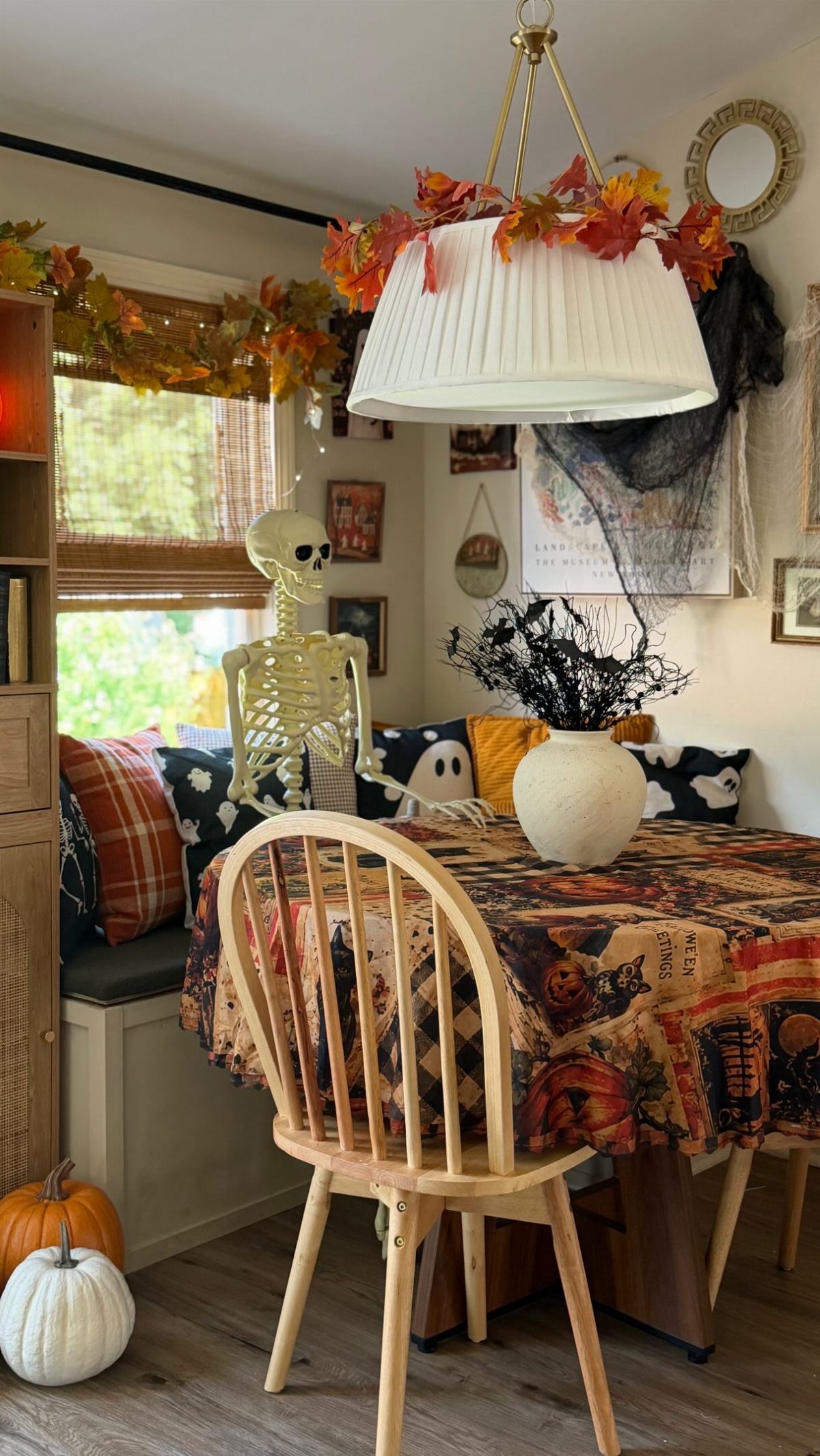 A Halloween-themed dining nook with a skeleton on a bench, patterned tablecloth, and autumn leaf garlands.