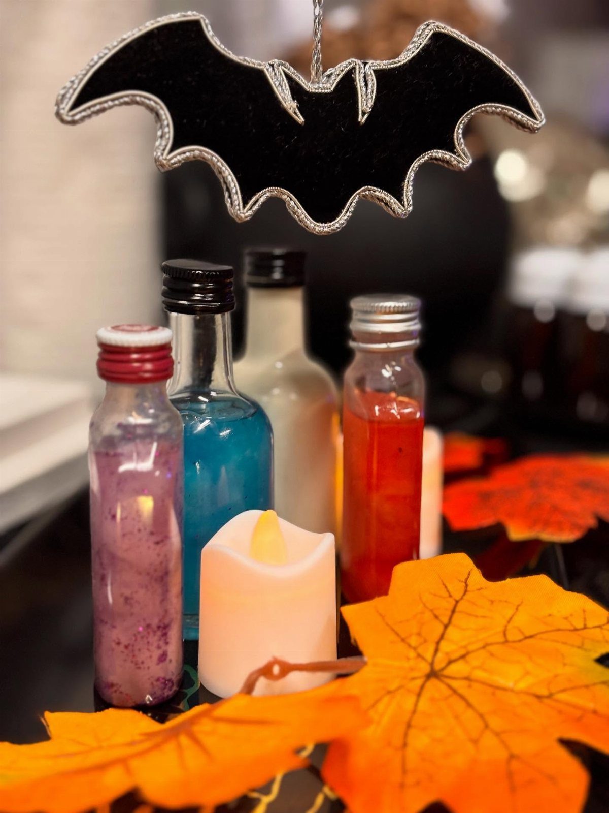 Small colorful potion bottles arranged with autumn leaves and a candle on a dark surface.