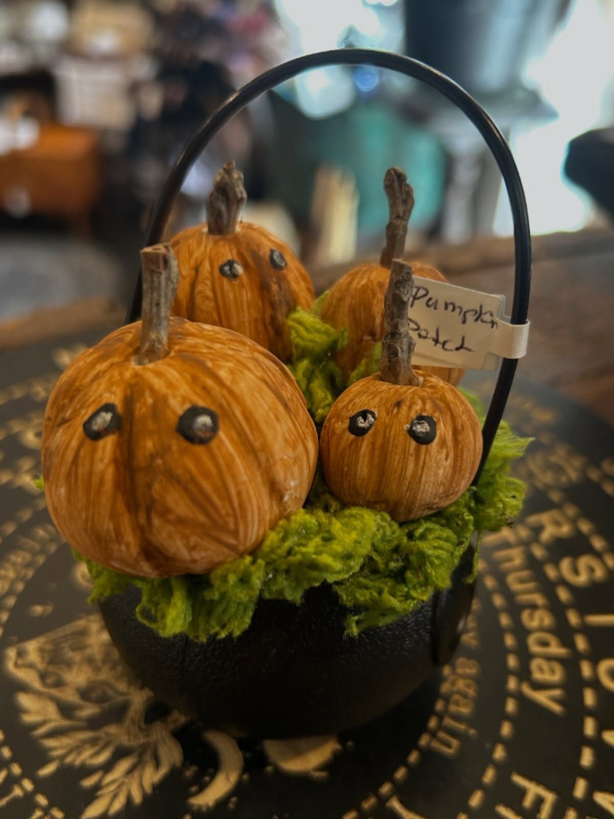 Four small orange pumpkins with black eyes resting in green moss inside a black cauldron.