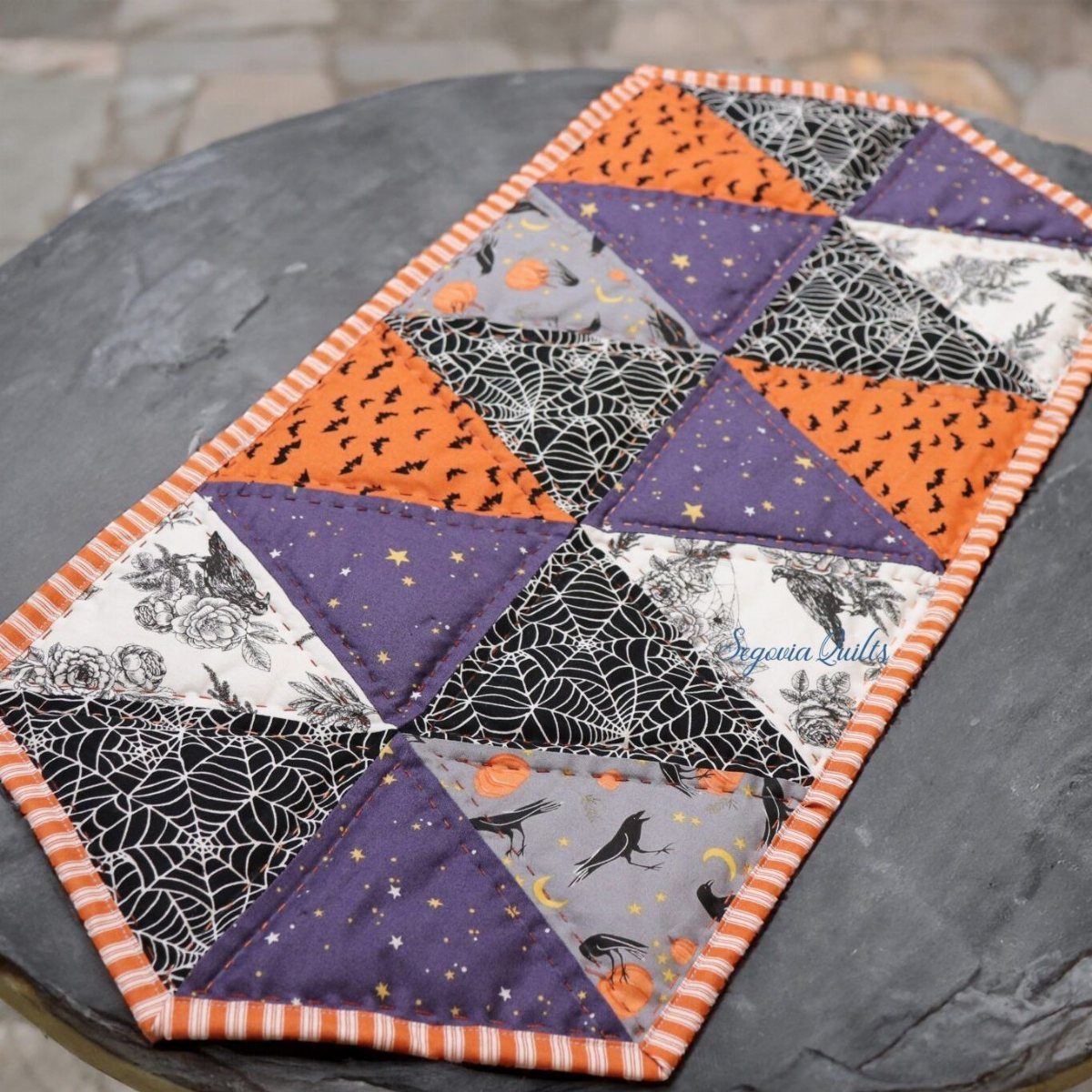 A patchwork quilted Halloween table runner with spiderweb, bat, and star patterns in orange, purple, and white.