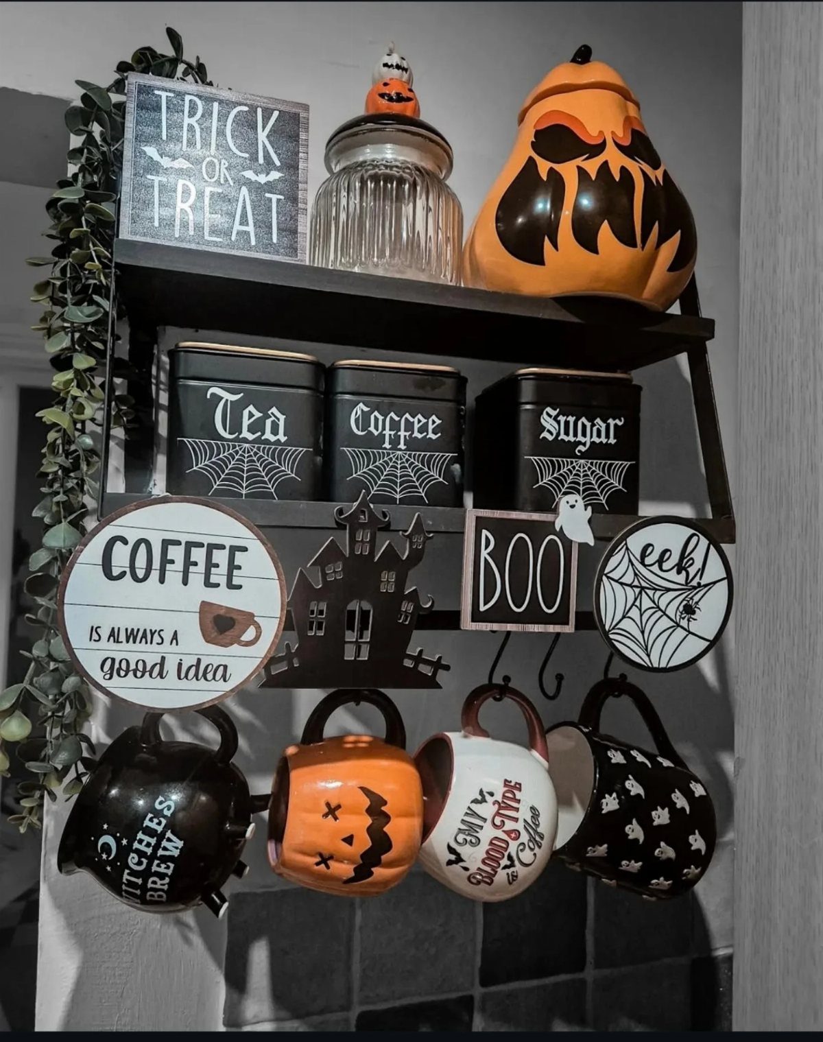 Halloween coffee station with black tea, coffee, and sugar canisters, jack-o'-lantern decor, and spooky hanging mugs.