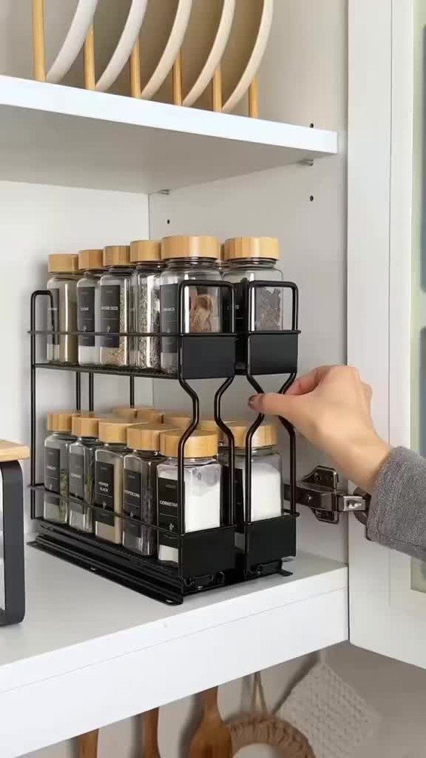 A hand reaching for glass spice jars organized in a black metal rack on a white shelf.